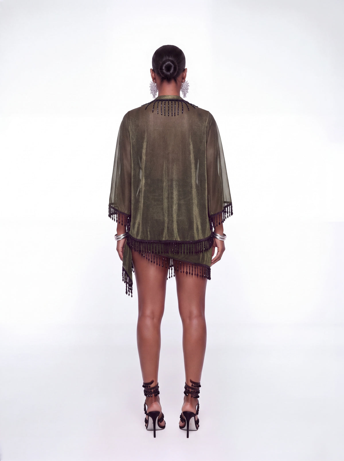 A model viewed from the back wearing a sage green velvet poncho-style cover-up with black fringe trim, paired with black lace-up high heels against a white studio background.