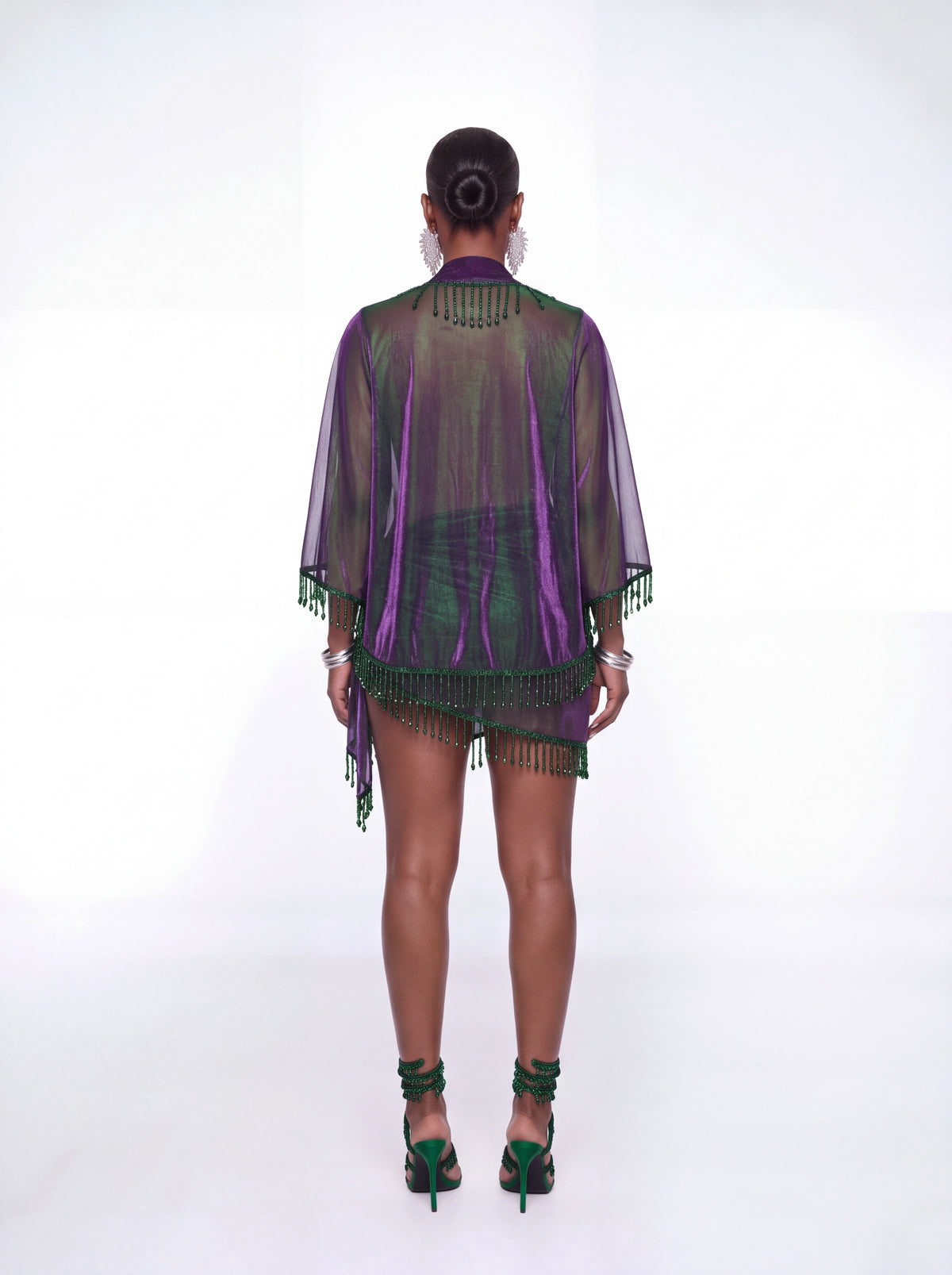 A model viewed from the back wearing a sheer purple and green fringe kimono cover-up over a matching purple and green mini skirt, paired with green high heels, on a white studio background.