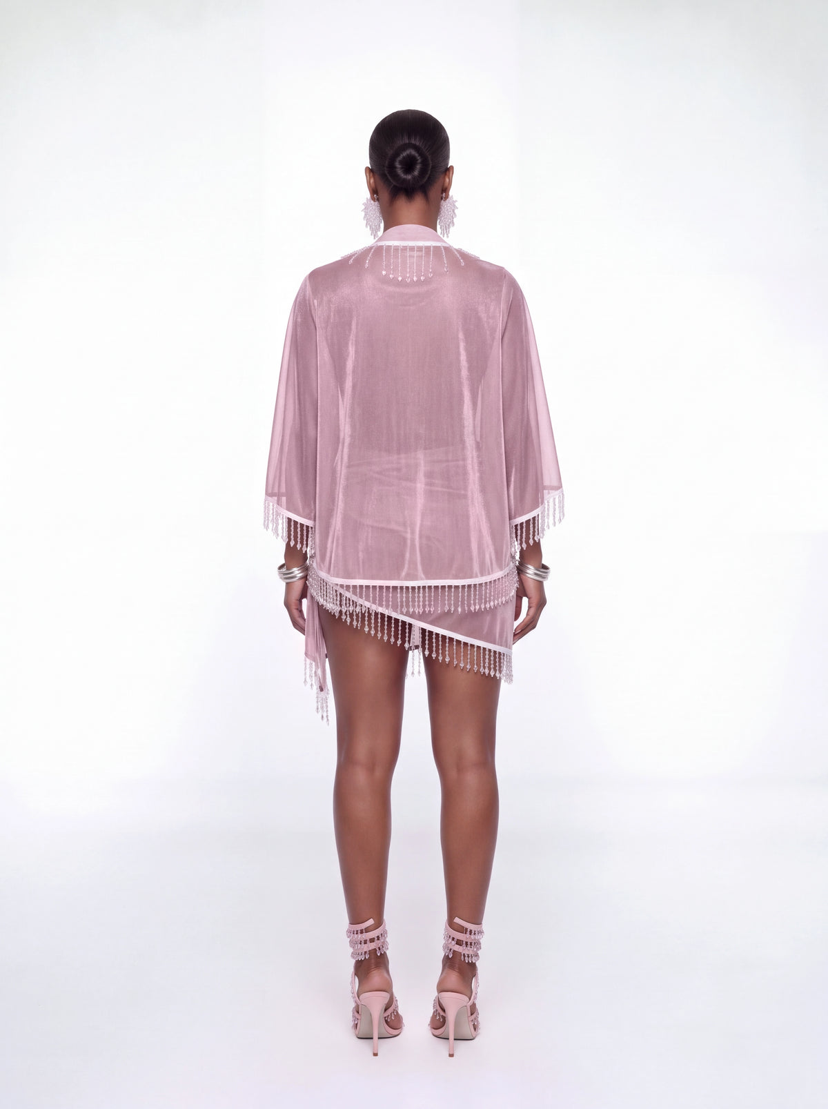 A model viewed from the back wearing a light pink sheer mesh cover-up with white fringe trim, paired with a matching pink mini skirt and pink high-heeled sandals, set against a plain white studio background.