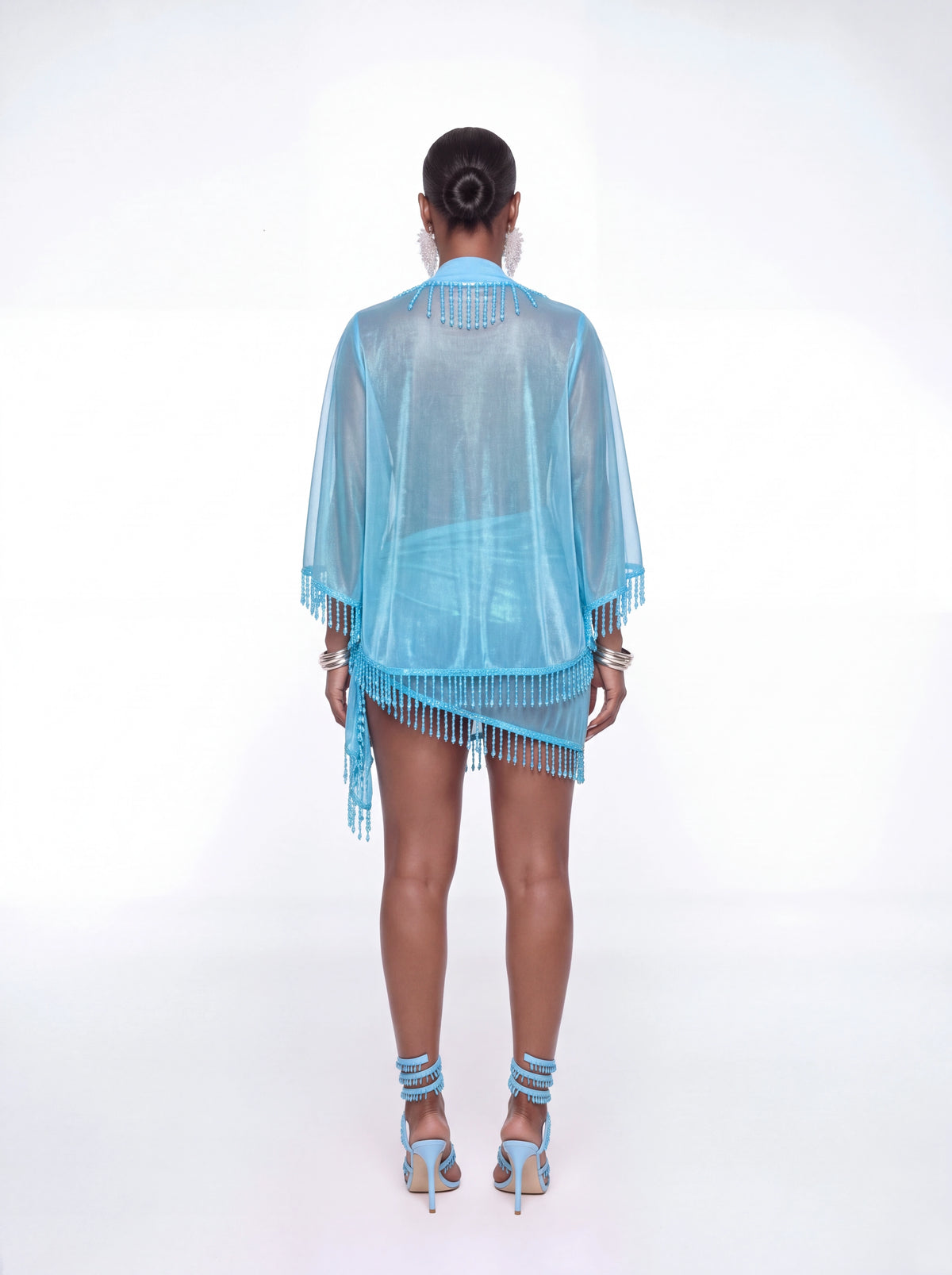 A model shown from the back wearing a light blue sheer fringe kimono cover-up with matching blue strappy high heels against a white studio background.