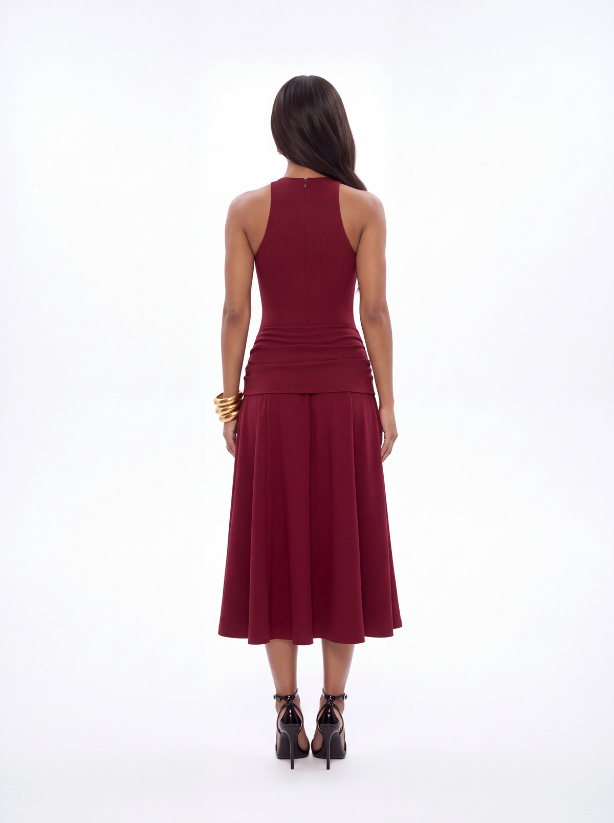 A woman viewed from the back wearing a sleeveless, high-neck, midi-length pleated dress in deep burgundy, paired with black lace-up heels and gold bracelets, against a plain white studio background.