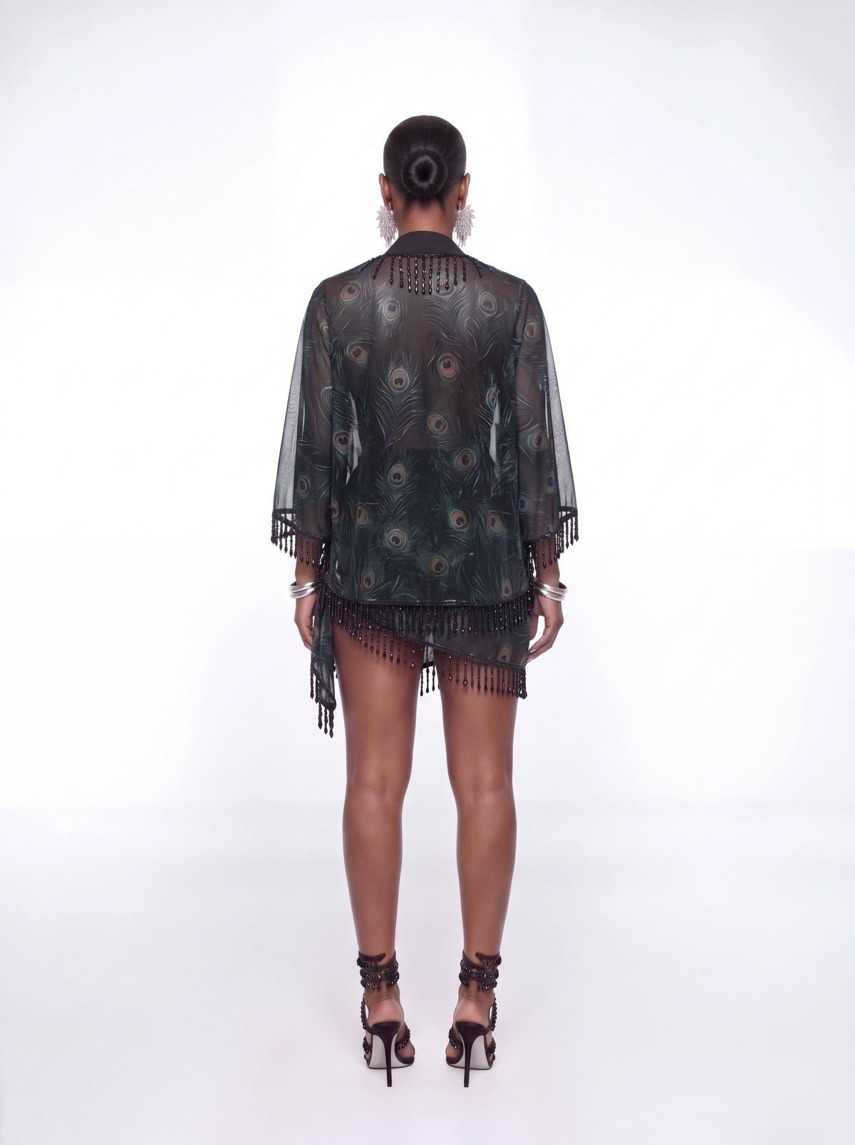 A woman viewed from the back wearing a sheer black fringed cover-up with a peacock feather print, paired with black strappy high heels, against a white studio background.