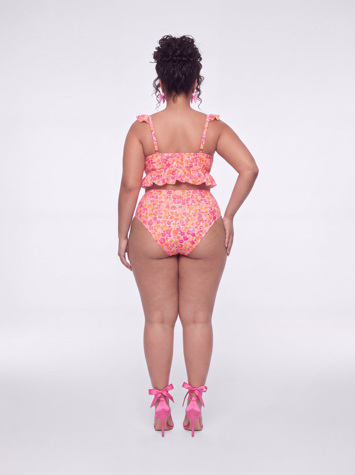 A model shown from the back wearing a pink and orange floral print one-piece swimsuit with ruffle straps, paired with pink high heels, against a light gray studio background.