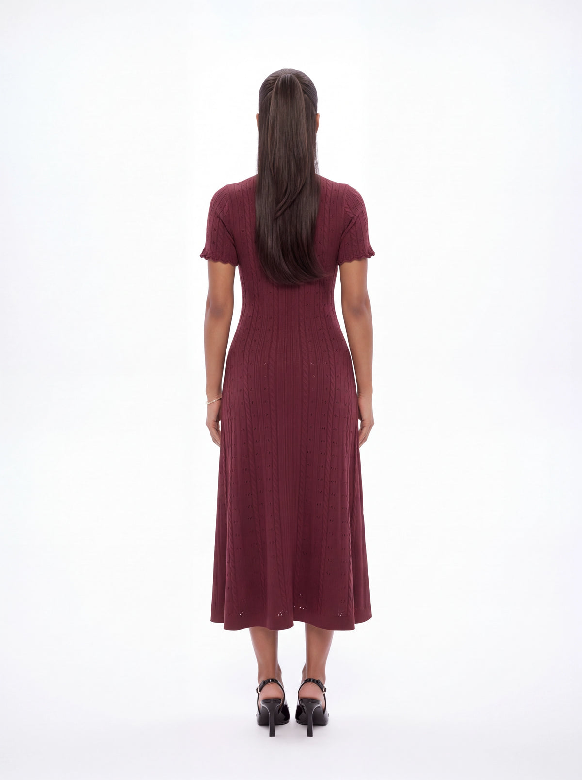 A woman viewed from behind wearing a short-sleeve, ribbed midi dress in a deep burgundy or maroon color, paired with black heels, against a plain white studio background.