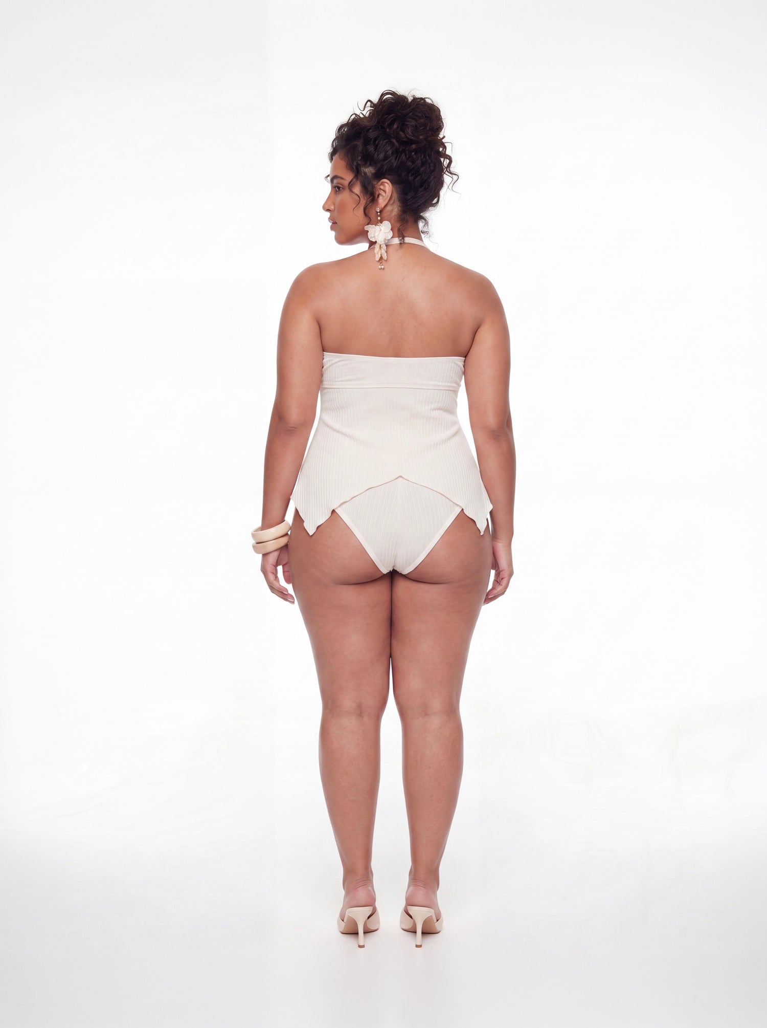 Back view of a cream ribbed one-piece swimsuit featuring a strapless back design, pointed handkerchief hem, and moderate coverage bottom. Styled with statement floral earrings and neutral heels against a white studio background.