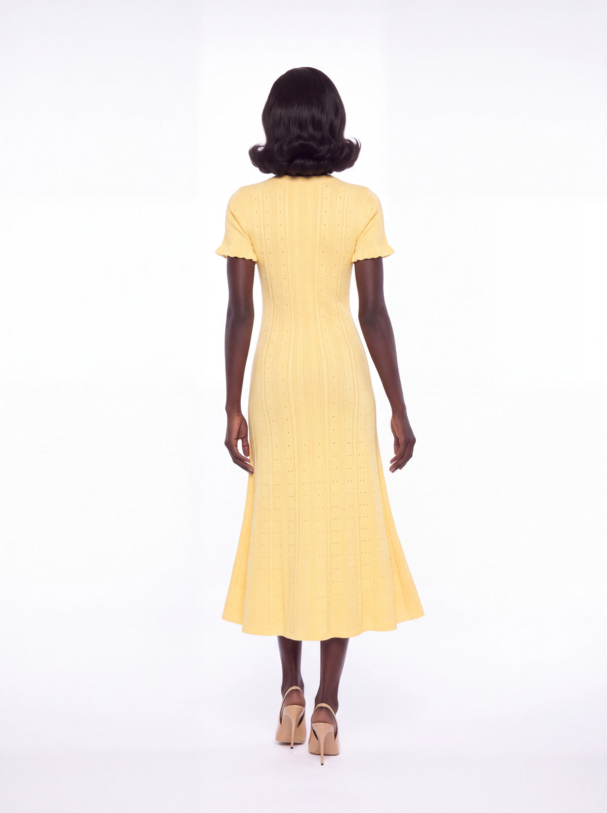 A woman viewed from behind wearing a pale yellow short-sleeve midi dress with a textured vertical stripe pattern, paired with beige high heels, against a solid white studio background.