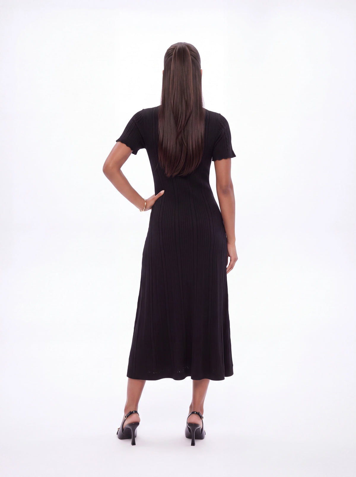 A woman viewed from behind wearing a short-sleeve black midi dress with a pleated skirt and black high heels, standing against a plain white studio background.