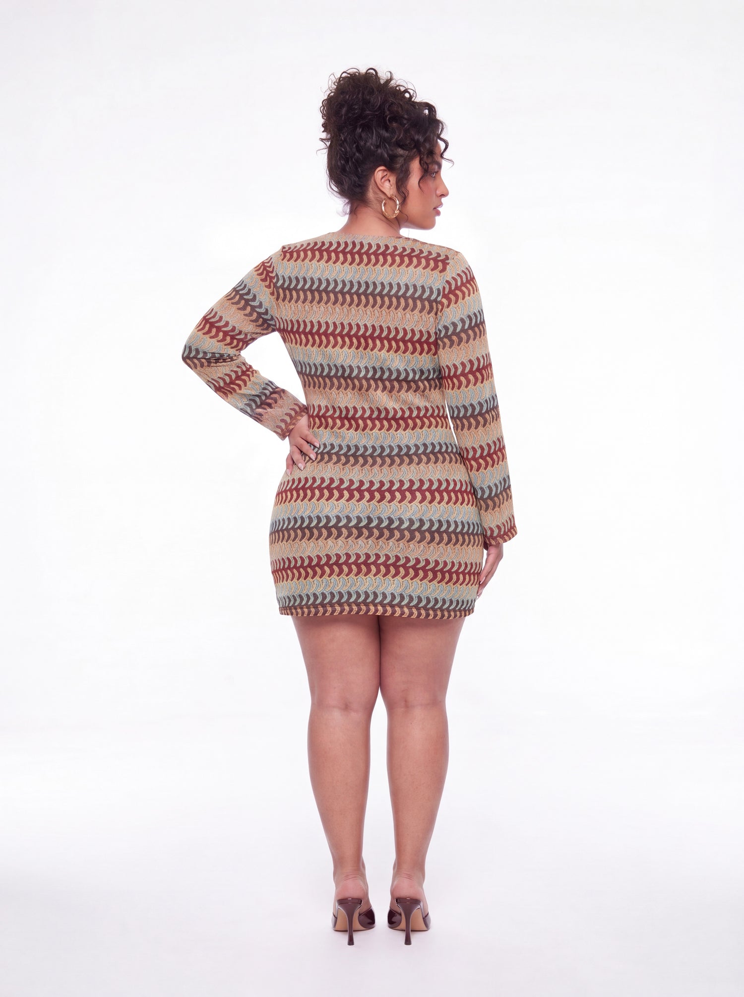 Back view of a fitted long-sleeve mini dress featuring a multicolor wave print, bodycon silhouette, and full-coverage back. Styled with gold hoop earrings and brown heels against a white studio background.