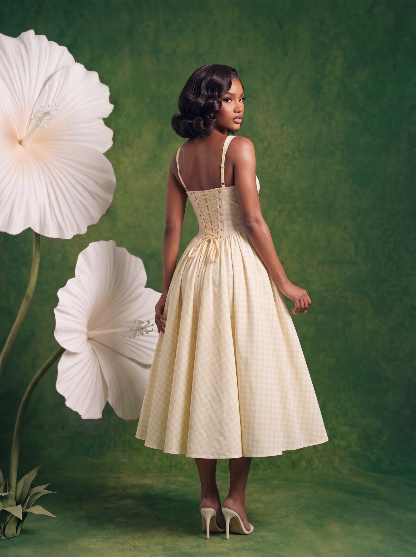 Back view of a butter-yellow gingham midi dress featuring adjustable shoulder straps, a lace-up corset back with gold grommet detailing, a cinched waist, and a flowing pleated midi skirt. Styled with white heeled mules against a green backdrop with oversized white flowers.