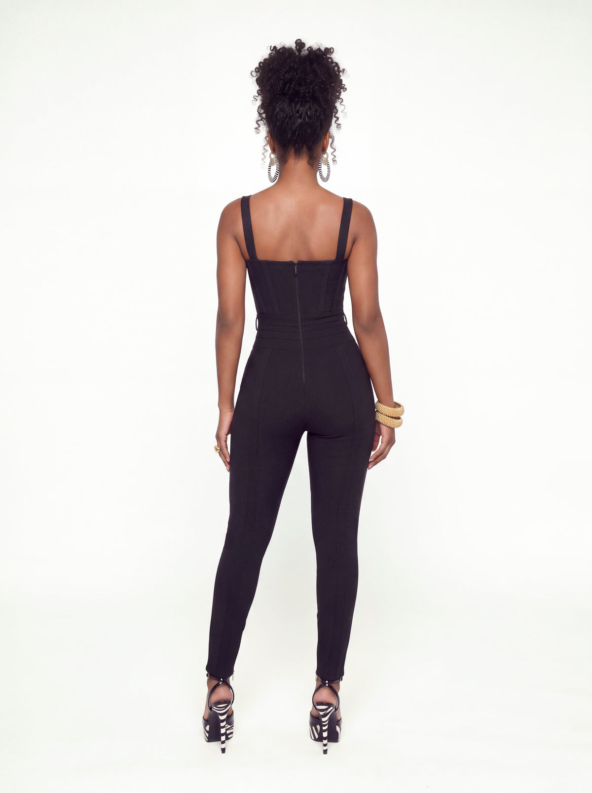 Back view of a black ribbed, form-fitting jumpsuit featuring wide shoulder straps, a straight back neckline, a visible center-back zipper, a fitted waist, and slim, tapered pant legs.