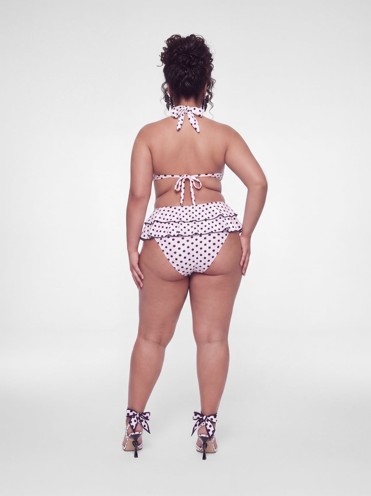 A woman stands with her back to the camera wearing a white two-piece bikini with black polka dots, featuring a ruffled halter top and matching ruffled bikini bottoms, paired with patterned high heels on a light gray studio background.