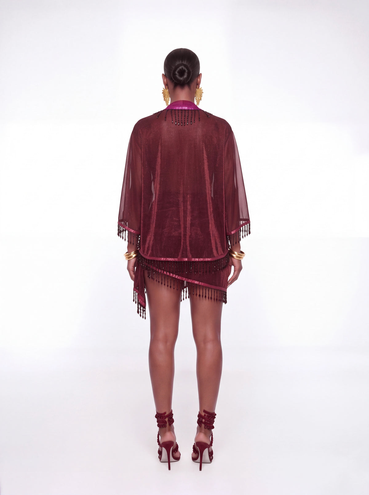 A model shown from the back wearing a sheer burgundy velvet cover-up with fringe details, paired with matching burgundy high-heeled sandals and gold jewelry, set against a plain white studio background.