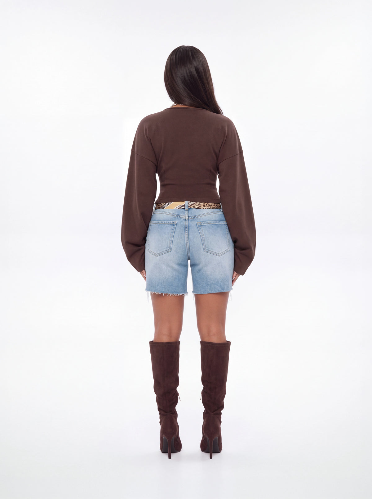 A woman is shown from behind wearing a brown long-sleeve cropped top, light blue denim shorts, and dark brown knee-high heeled boots against a plain white studio background.