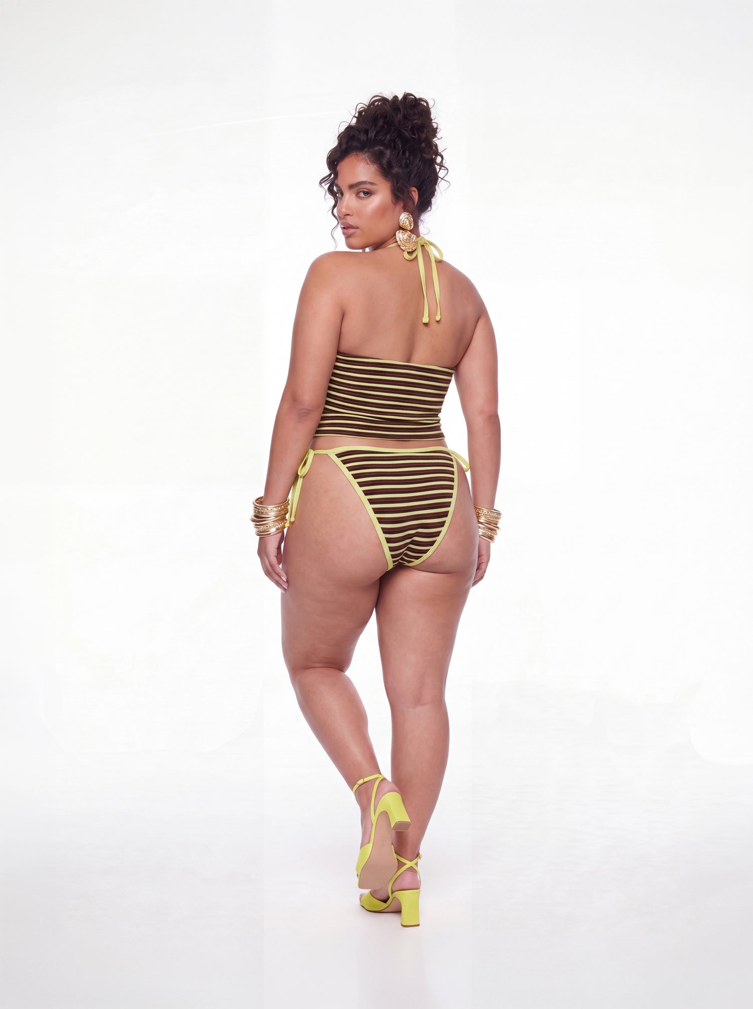 Back view of a brown and yellow striped tankini swim set featuring a halter tie closure, fitted tankini top, and matching side-tie bikini bottoms. Styled with gold statement earrings, stacked bangles, and yellow heels against a white studio background.