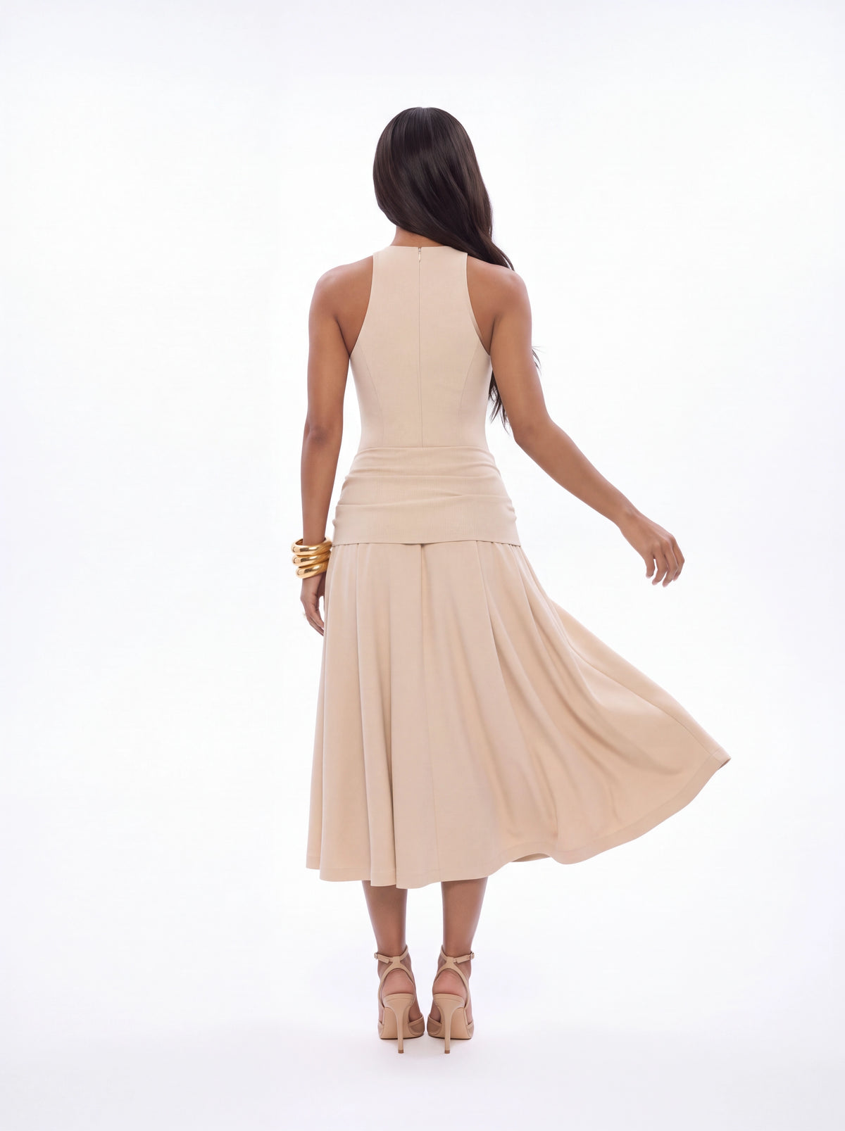 A woman shown from behind wearing a sleeveless beige midi dress with a gathered waist and flowing skirt, paired with beige high-heeled sandals and gold bracelets, against a plain white studio background.