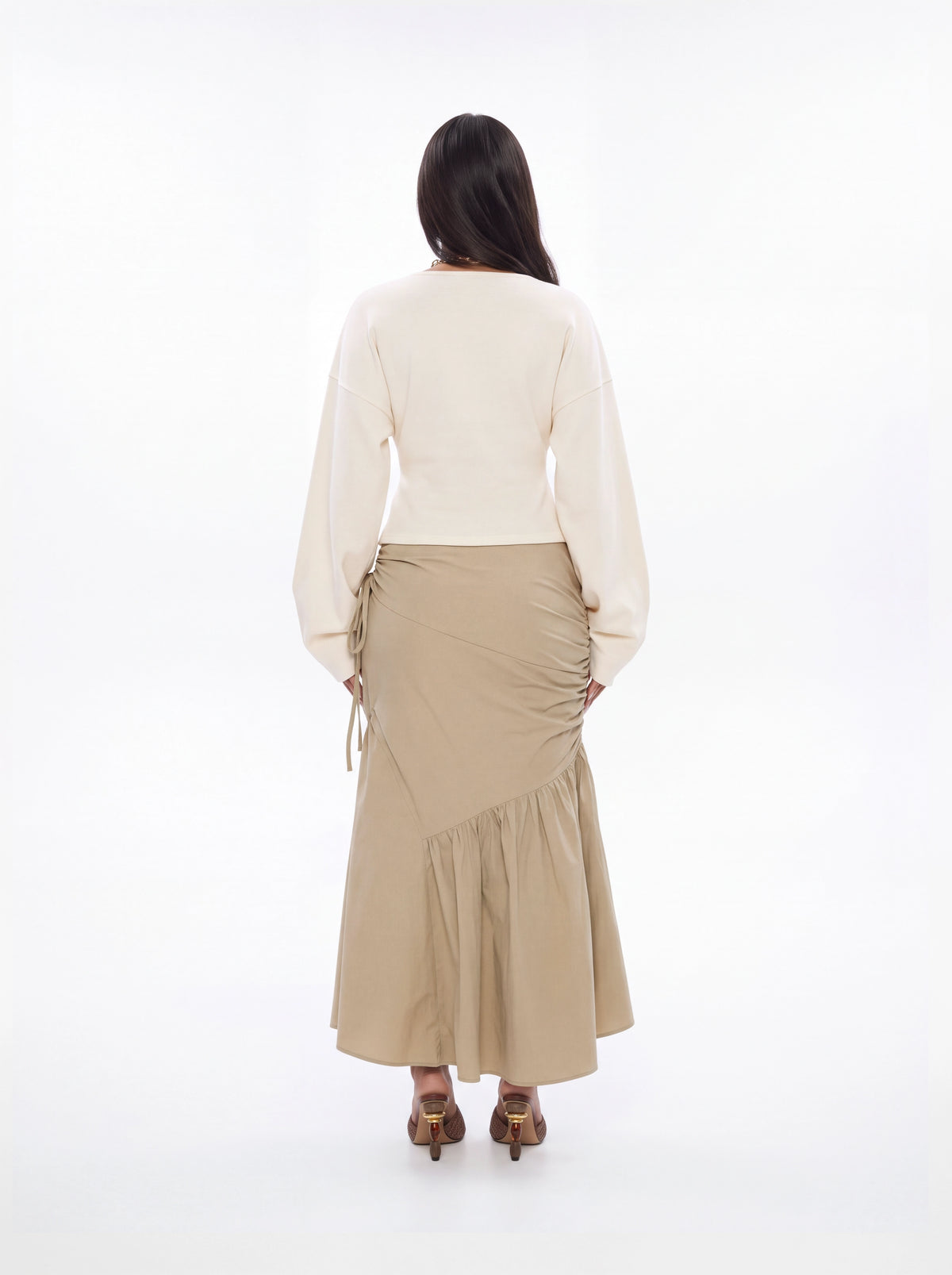A woman viewed from behind wearing a cream long-sleeve top and a beige ruched wrap maxi skirt with side ties, paired with brown heels, against a solid white studio background.