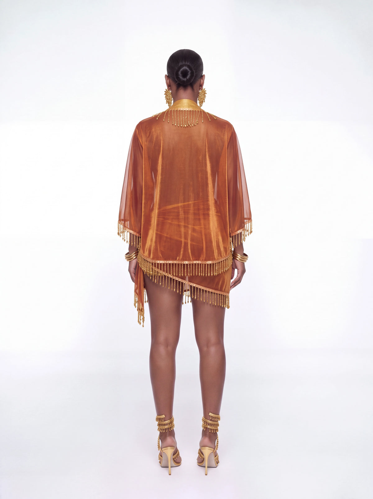 A model is shown from the back wearing a sheer, metallic bronze-orange fringed cover-up dress with gold accessories, set against a plain white studio background.
