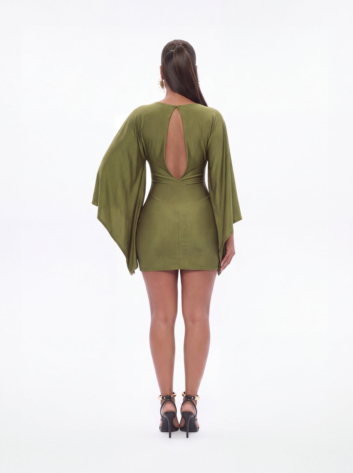 A woman viewed from the back wearing a short olive green dress with wide draped sleeves and a large open back, paired with black high-heeled sandals against a plain white studio background.