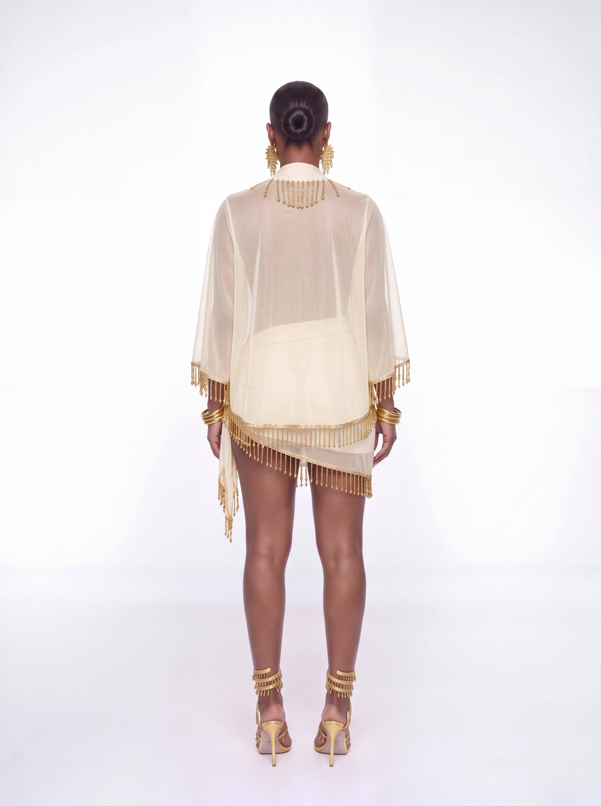 A model shown from the back wearing a sheer cream-colored fringed cover-up and matching mini skirt, styled with gold jewelry and heels against a white studio background.