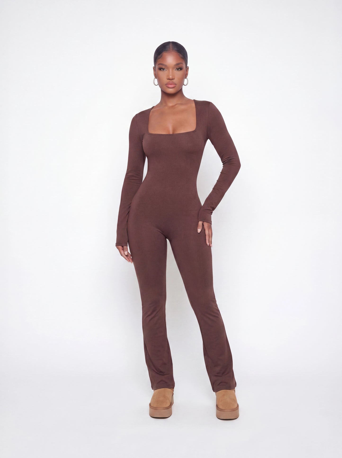 Model wearing a brown long sleeve ribbed jumpsuit with a square neckline, curve-hugging fit, and full-length silhouette in soft stretchy fabric.