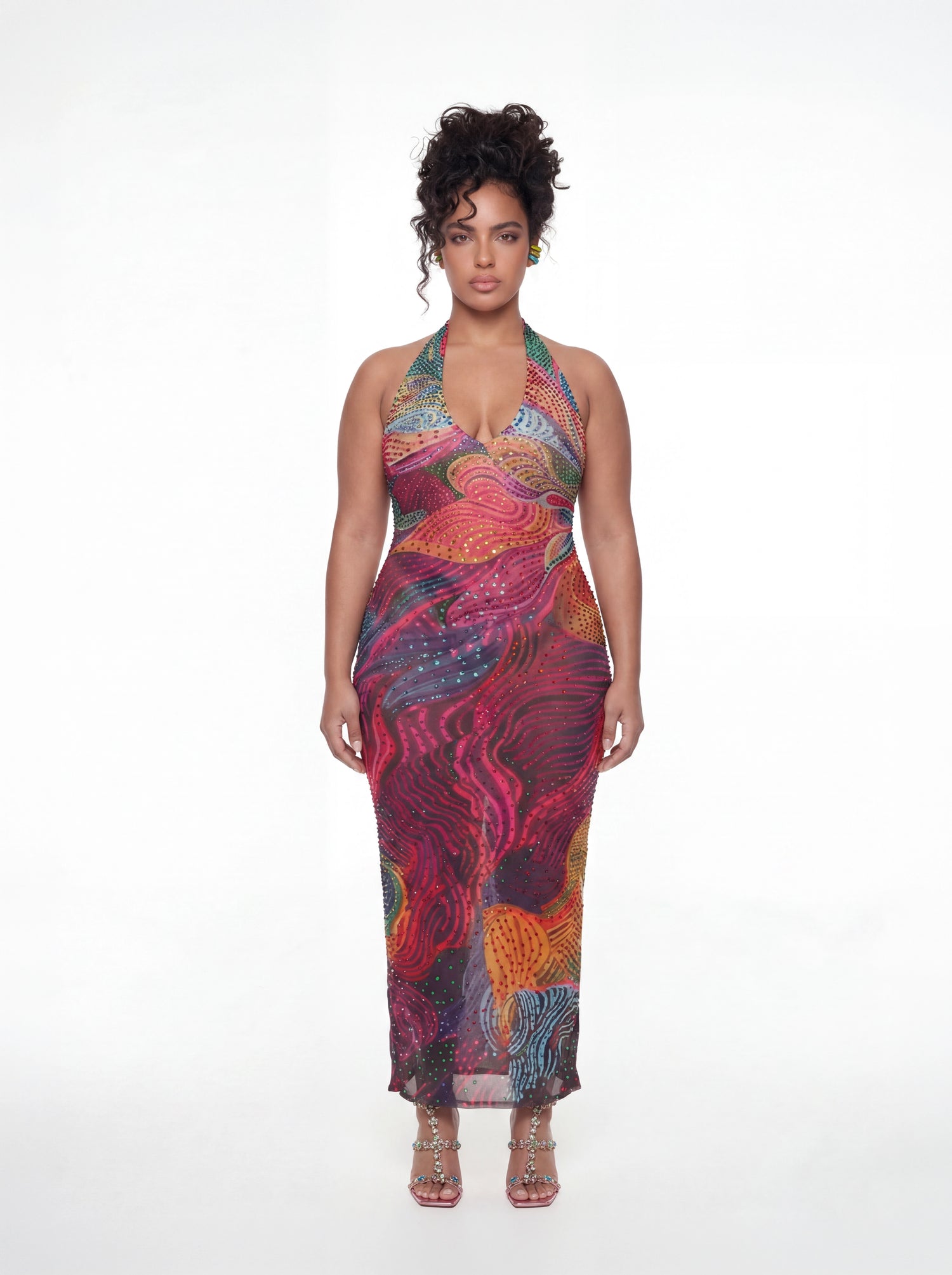 Curve model wearing a colorful abstract-print halter midi dress featuring a plunging neckline, fitted silhouette, and allover multicolor rhinestone embellishments. The dress showcases vibrant shades of pink, purple, orange, blue, and green with a swirling artistic pattern. Styled with jeweled strappy heels against a white studio background.