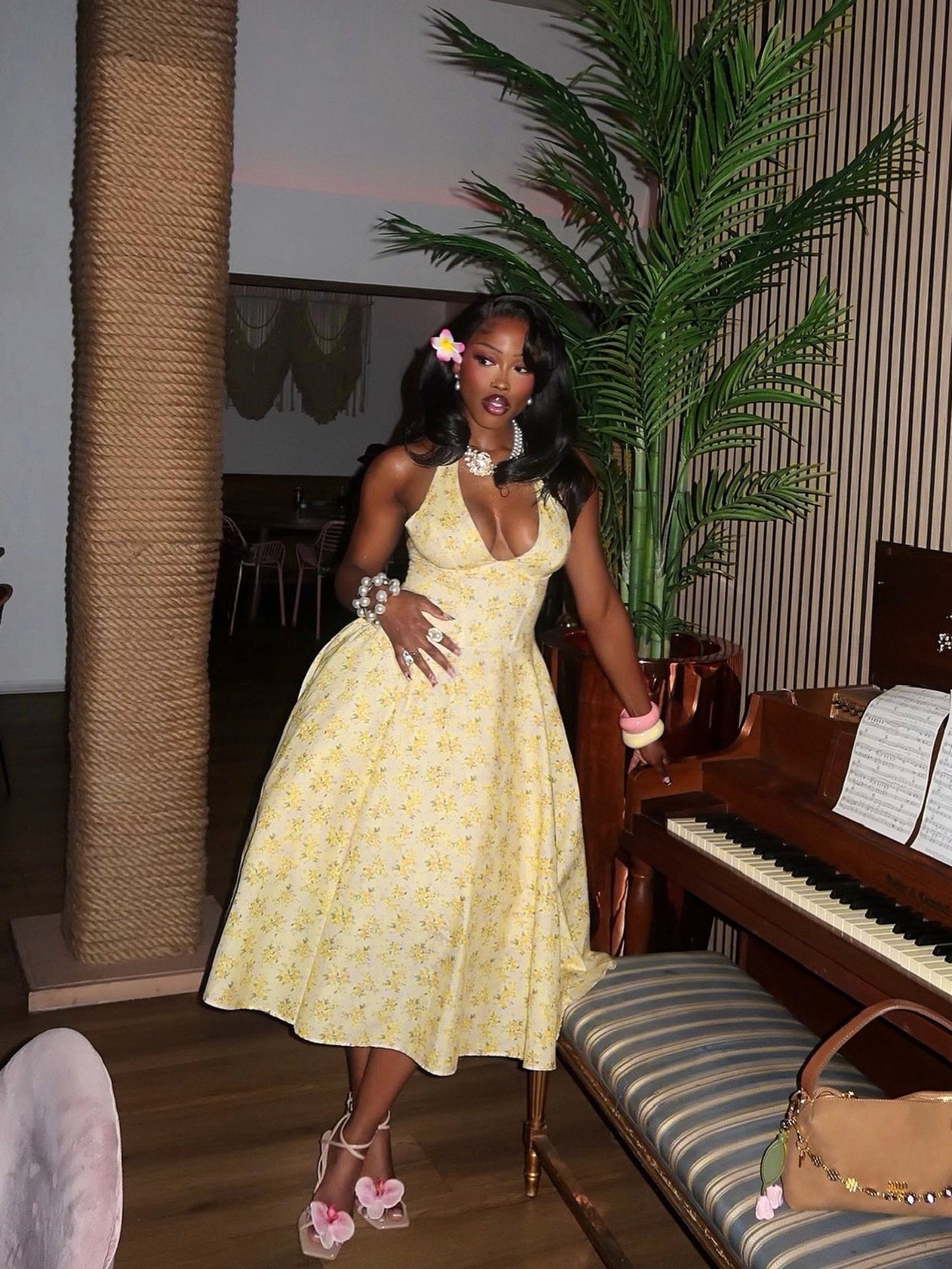  Model wearing a yellow floral halter midi dress with a fitted corset-style bodice and full A-line skirt. Styled with floral high heels, chunky pearl bracelets, statement rings, a crystal necklace, and a pink flower in her hair, posing beside a piano in a tropical-inspired indoor setting.