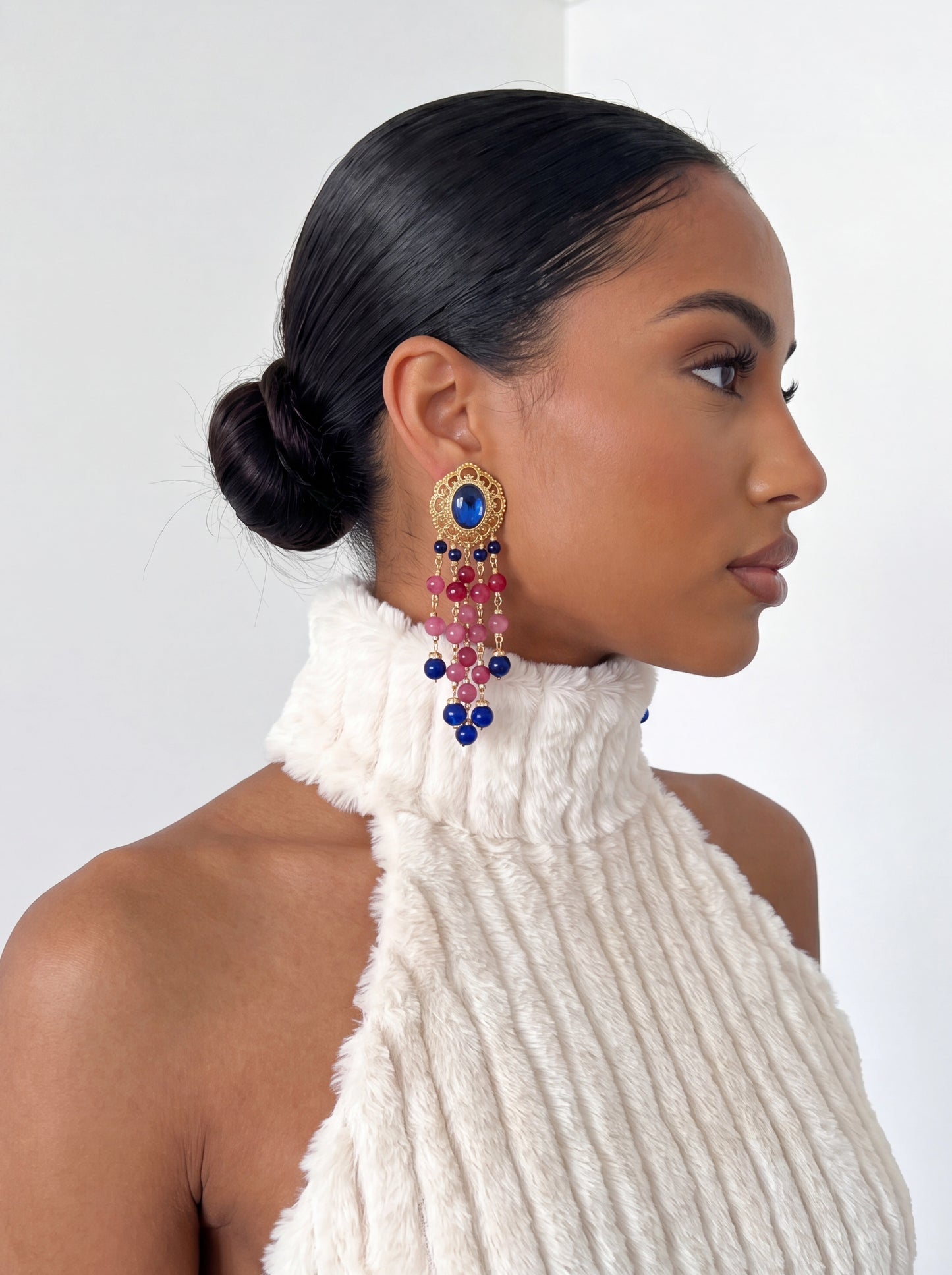 Side profile of a model wearing gold-tone statement earrings featuring a sapphire-blue gemstone stud and cascading pink and blue bead fringe. Styled with a sleek low bun and cream textured halter top against a white studio background.