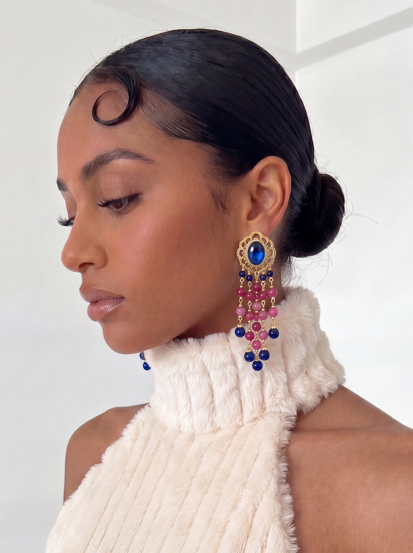 Close-up portrait of a model wearing vintage-inspired gold-tone chandelier earrings with a sapphire-blue center stone and dangling pink and blue beads. Styled with a sleek bun and cream textured halter neckline against a white background.