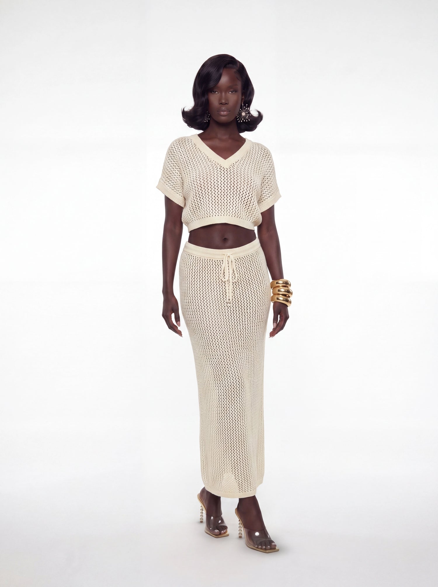 Model wearing a cream crochet two-piece set featuring a relaxed knit crop top with deep neckline and matching high-waisted maxi skirt. Styled with gold bangles and clear heels.