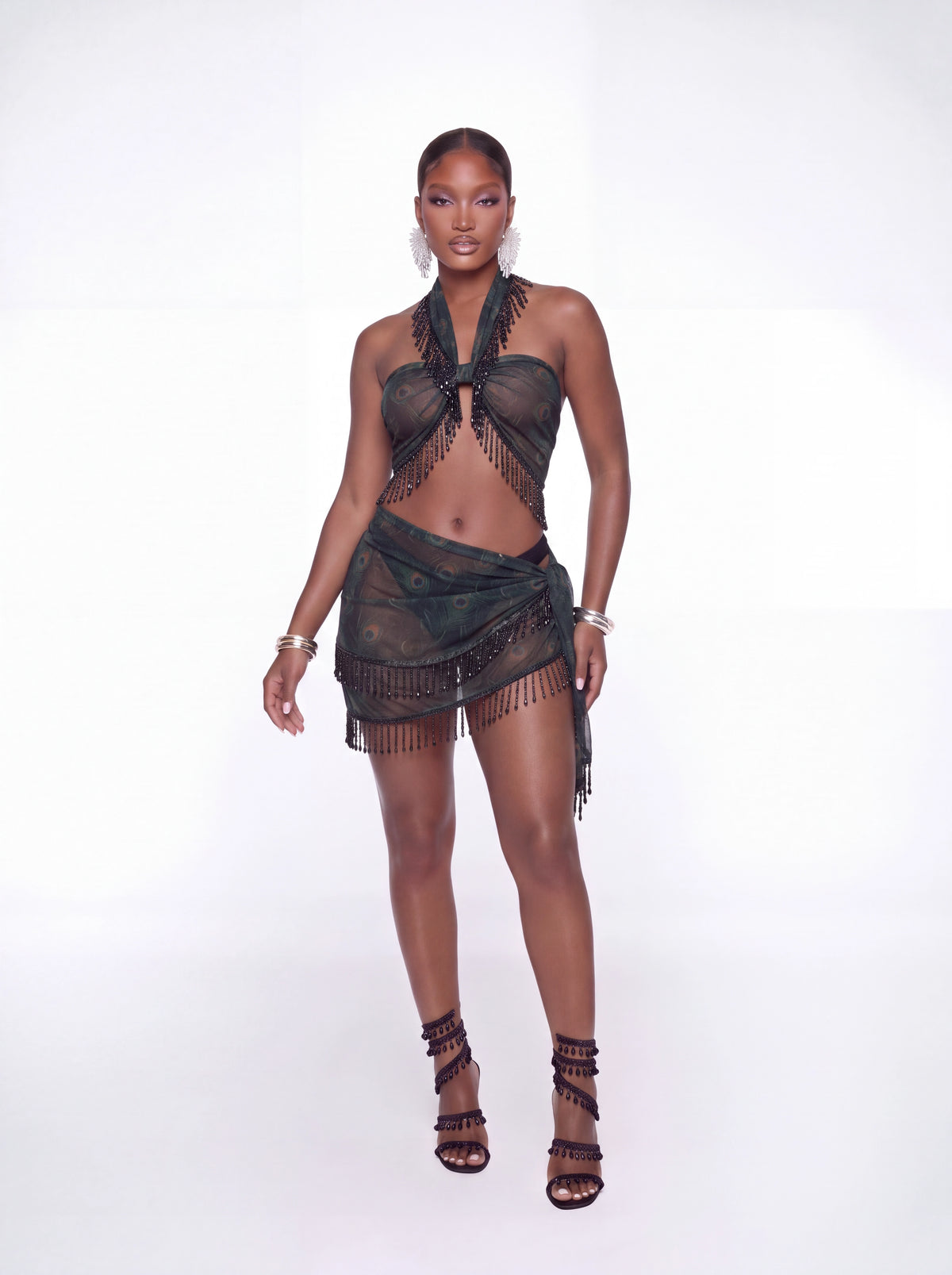 A model wearing a black sheer fringed halter bikini top and matching fringed sarong skirt, paired with black strappy high-heeled sandals, against a solid white studio background.