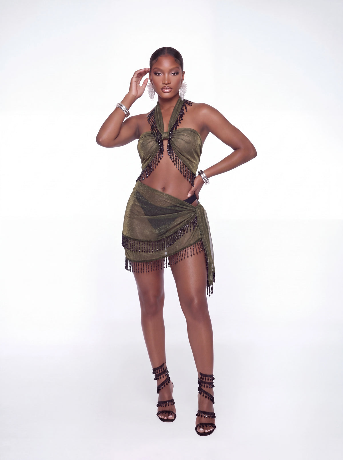 A model wearing a two-piece olive green and black fringed halter top and matching mini skirt, paired with black strappy high-heeled sandals, against a white studio background.