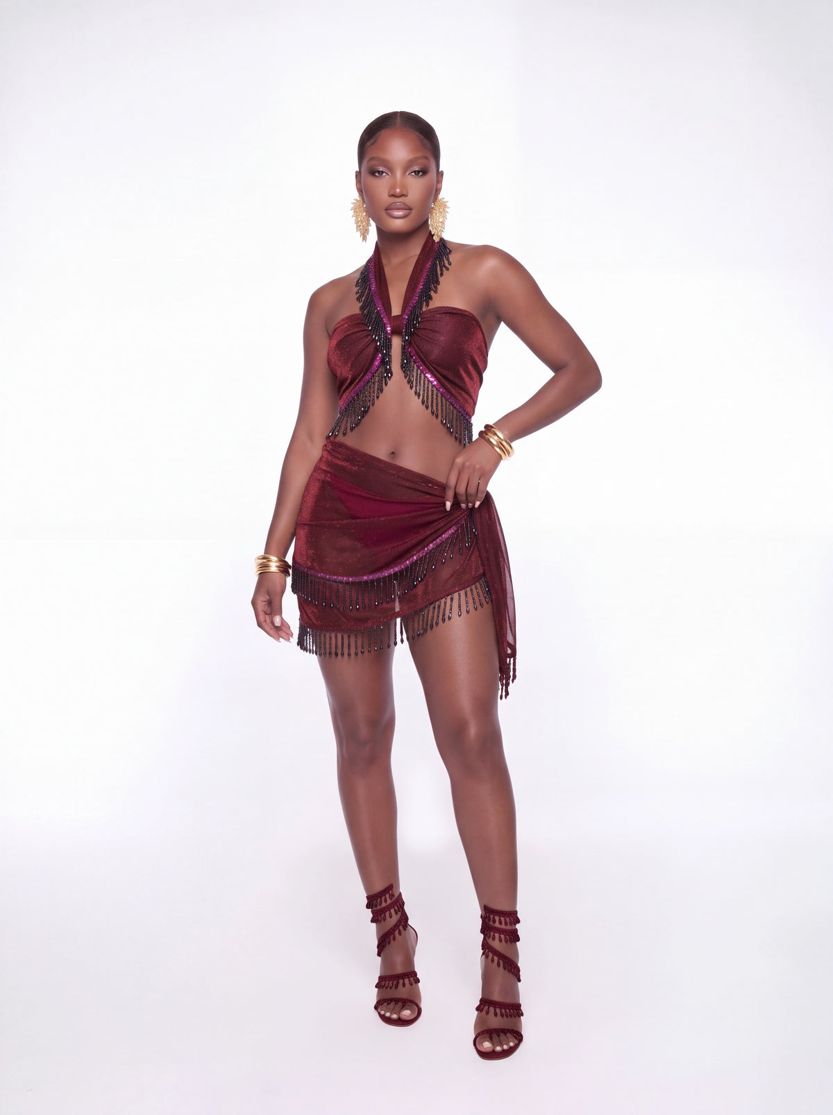 A model wearing a two-piece belly dance outfit in deep burgundy velvet with black fringe, paired with matching strappy high-heeled sandals and gold jewelry, against a plain white studio background.