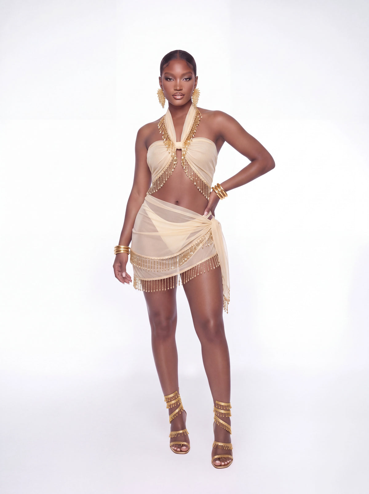 A model wearing a beige halter-neck scarf set with gold fringe and matching gold accessories, including earrings, bracelets, and strappy high-heeled sandals, posed against a solid white studio background.