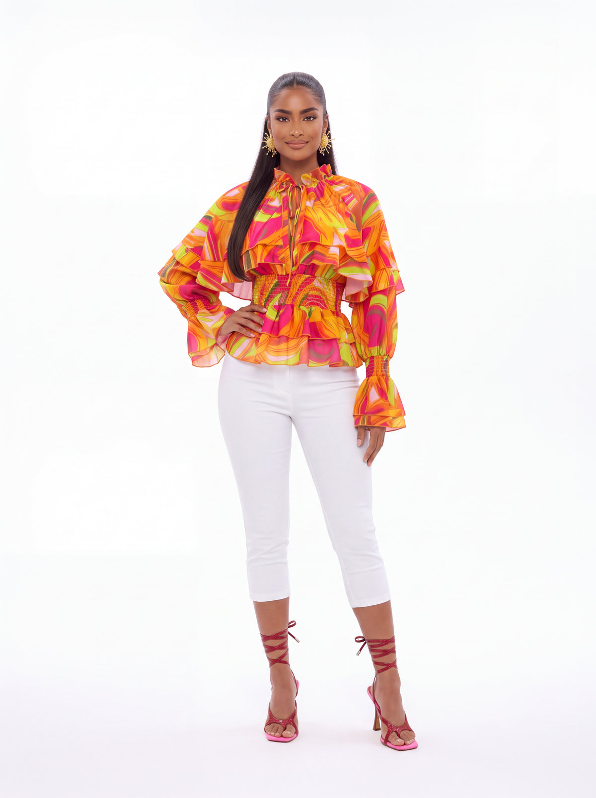 Model wearing the Vera Floral Top in coral, an orange and pink swirl print blouse with tiered ruffle sleeves, smocked waist and tie neck, styled with white capri pants.