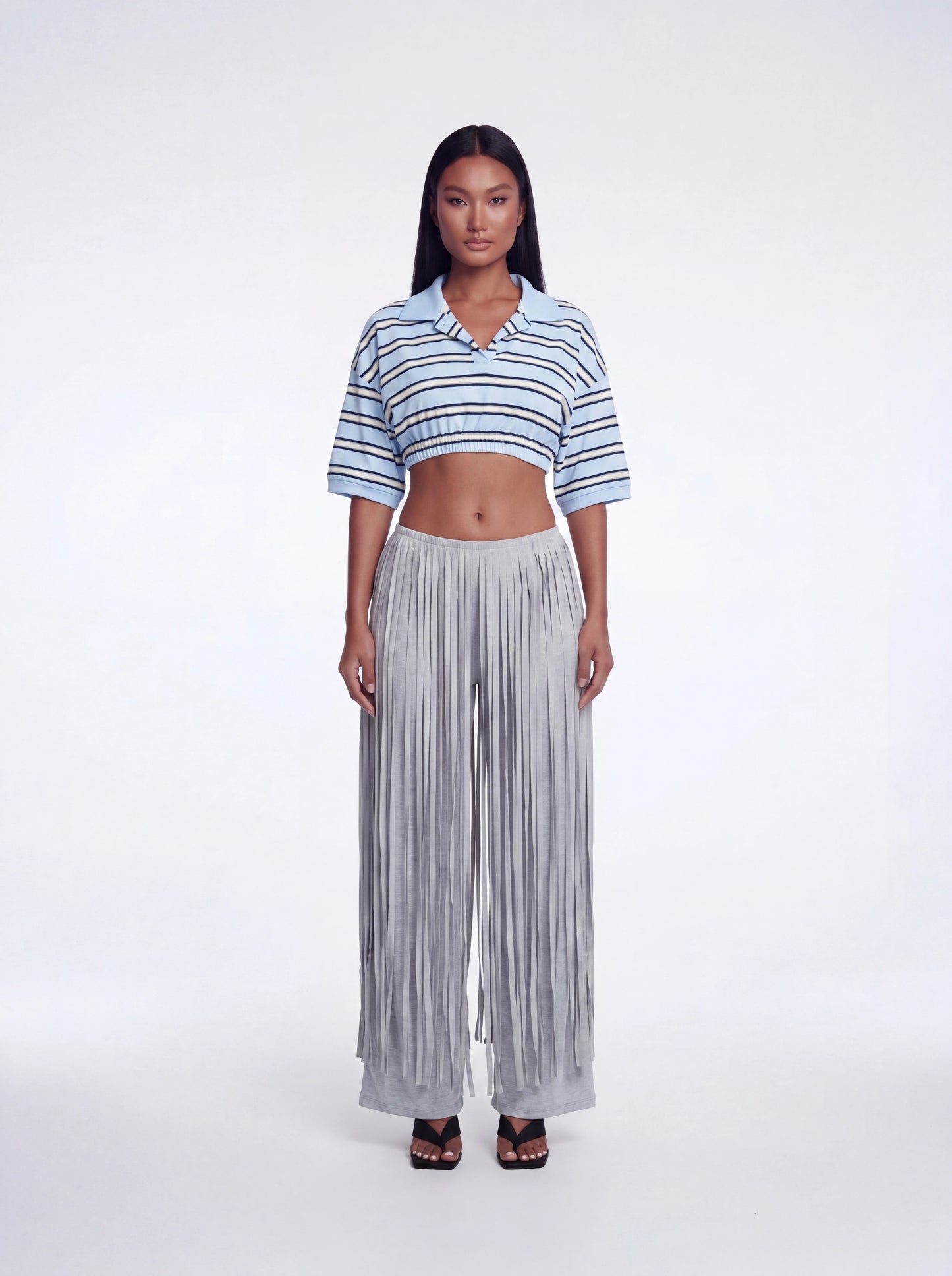 Model wearing grey high-waisted fringe pants with layered fringe detailing and a wide-leg fit paired with a blue striped cropped collared knit top and black heels.