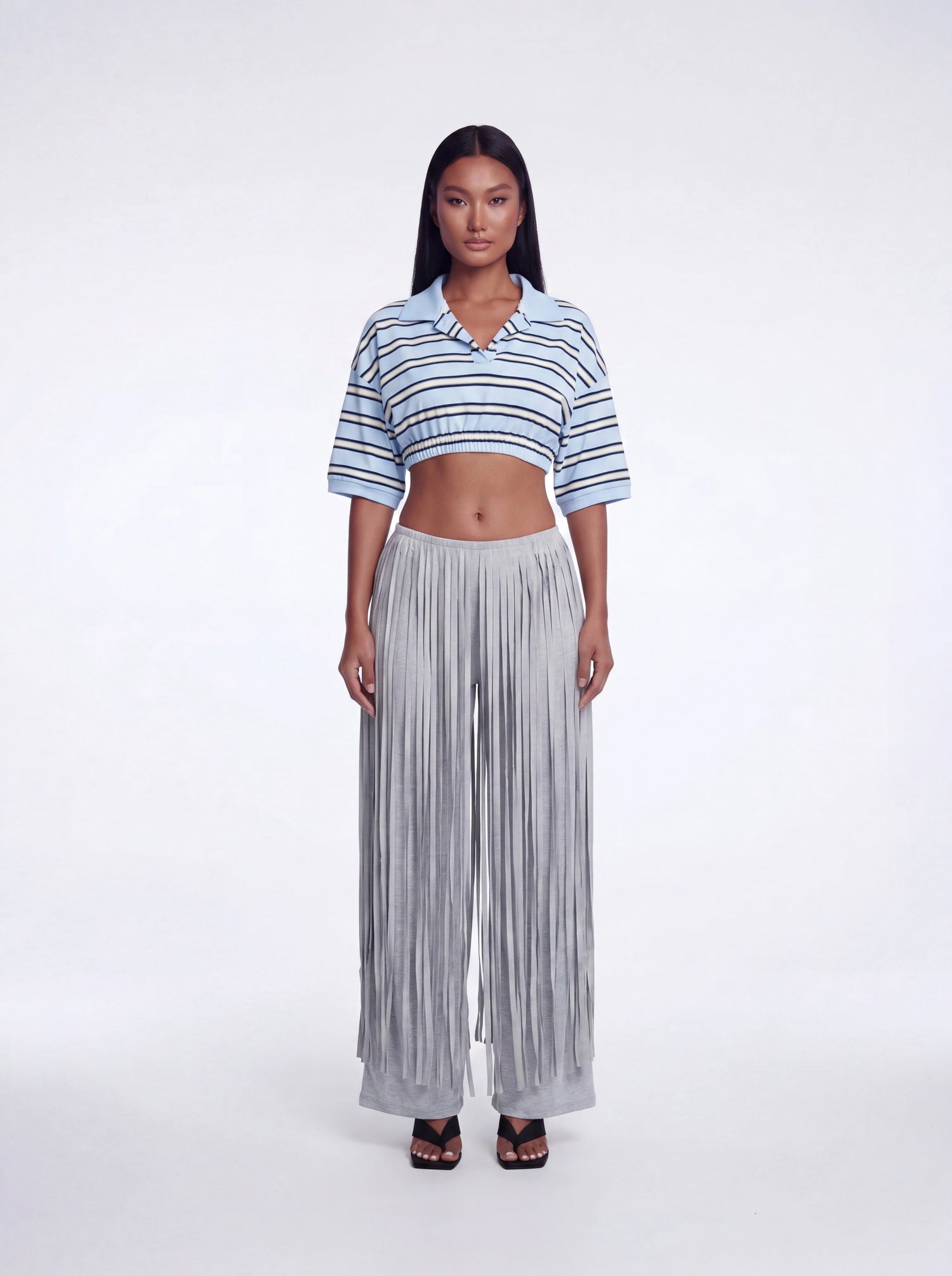 Model wearing grey high-waisted fringe pants with layered fringe detailing and a wide-leg fit paired with a blue striped cropped collared knit top and black heels.