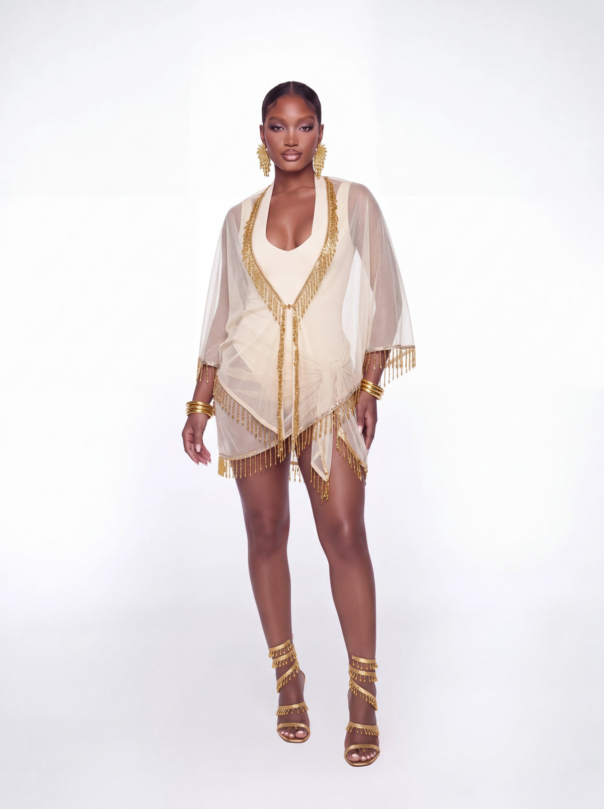 A model stands against a white studio background wearing a sheer cream kimono-style cover-up with gold fringe trim and matching gold accessories, including earrings, bracelets, and strappy high-heel sandals.