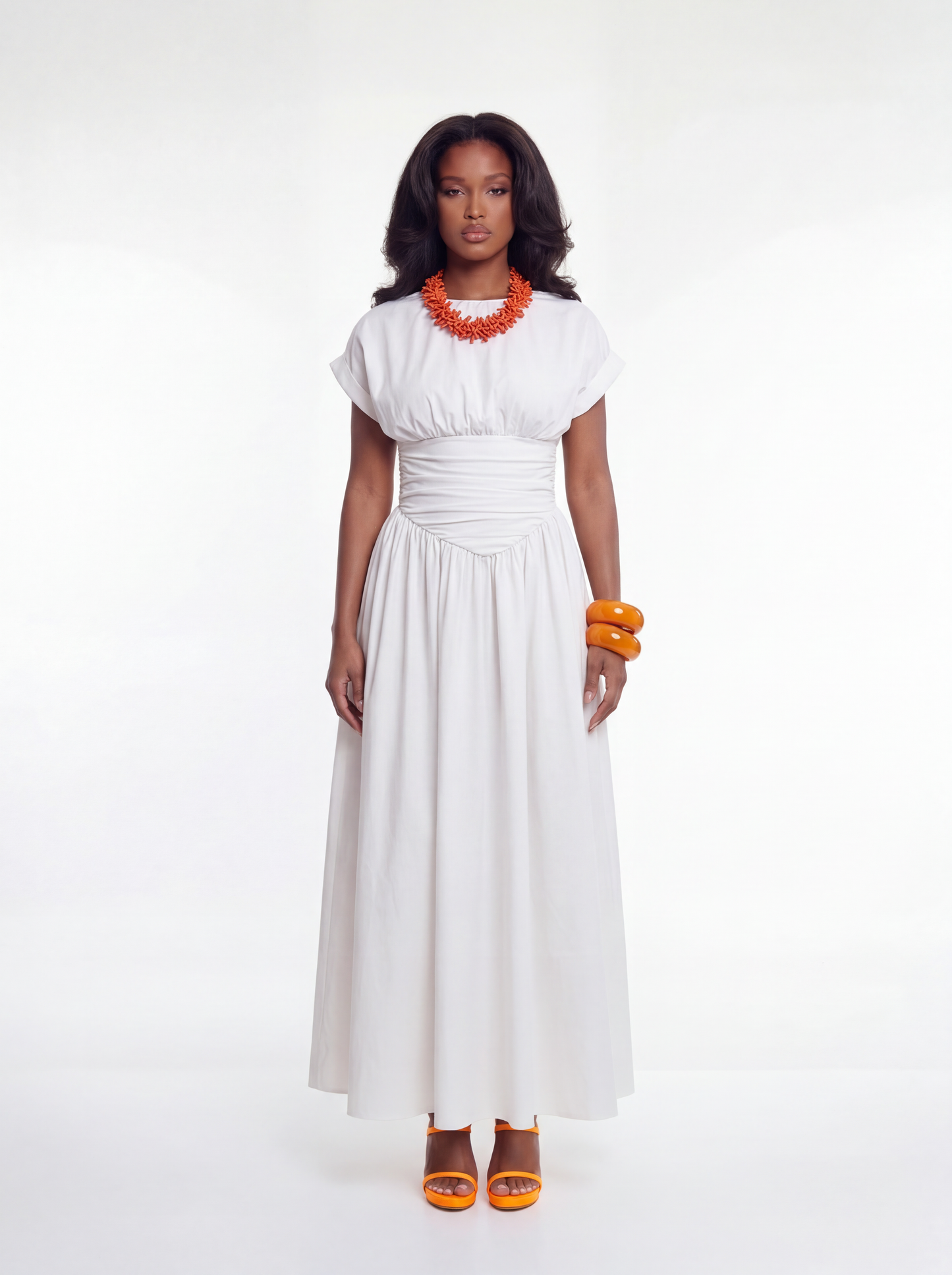 Woman wearing a white maxi dress with short sleeves and a ruched cinched waist, featuring a flowing full-length skirt, styled with an orange statement necklace, orange bangles, and matching heels.