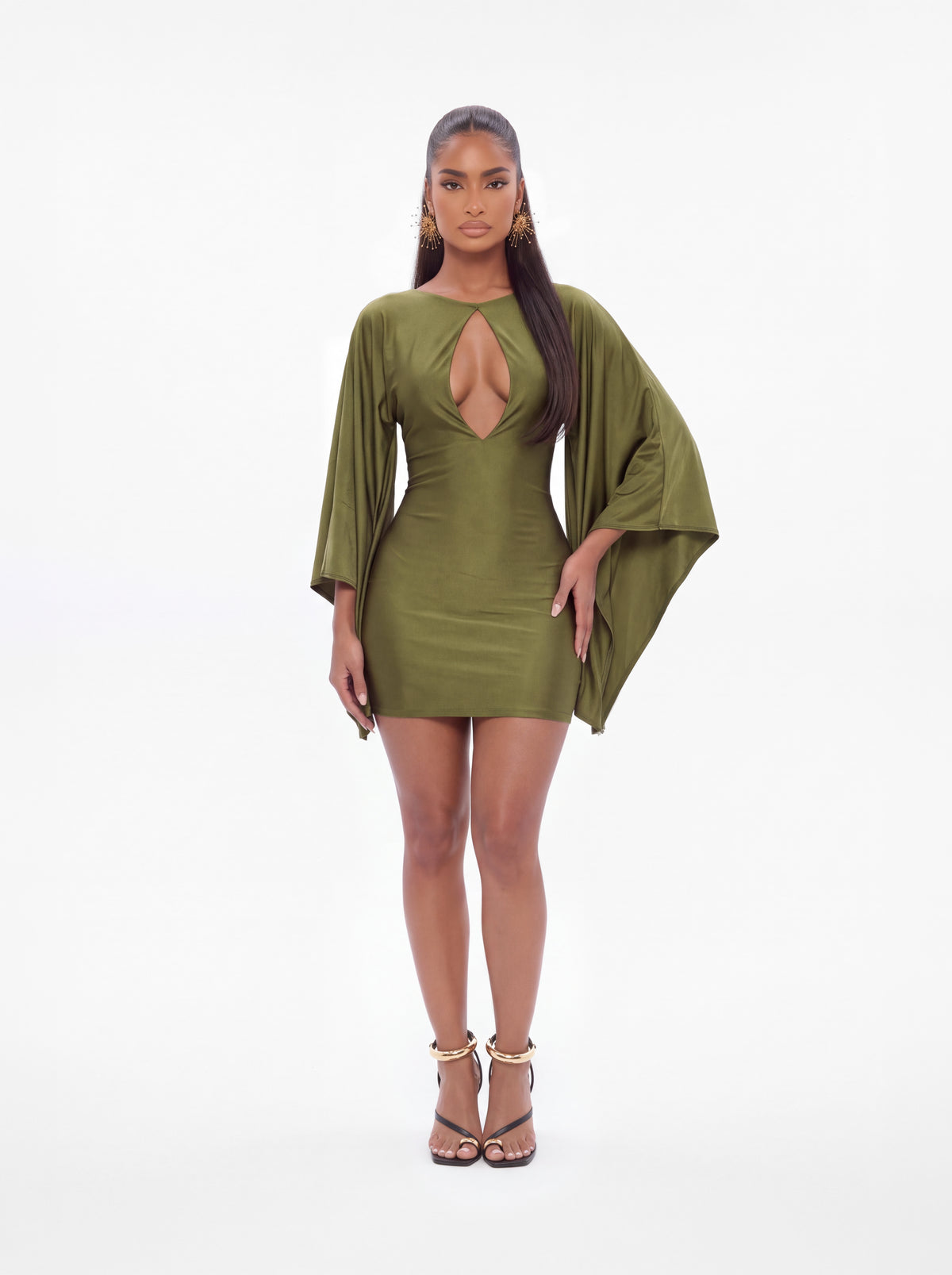 A woman modeling a short olive green bodycon dress with a plunging keyhole cutout at the chest, wide draped sleeves, and strappy high-heeled sandals, photographed against a plain white studio background.