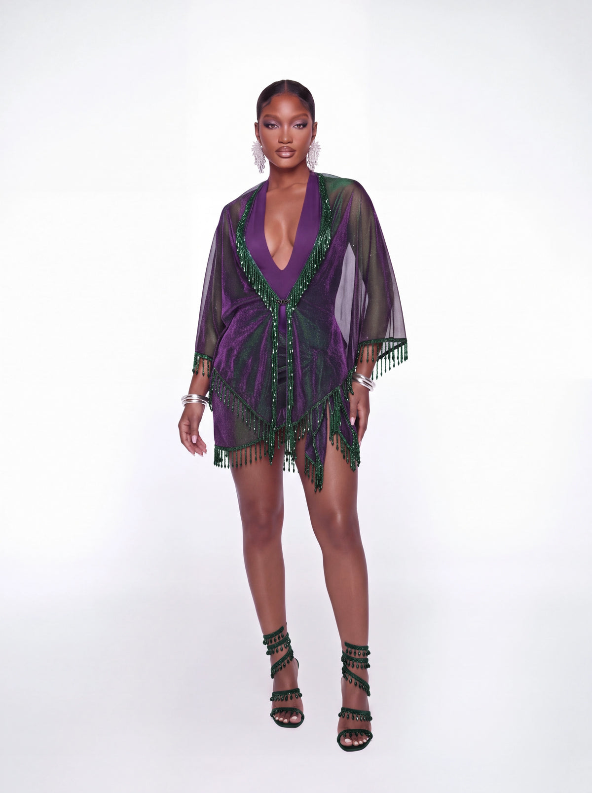 A model stands against a white studio background wearing a sheer purple and green beaded cover-up dress with fringe trim, paired with black strappy high-heeled sandals.