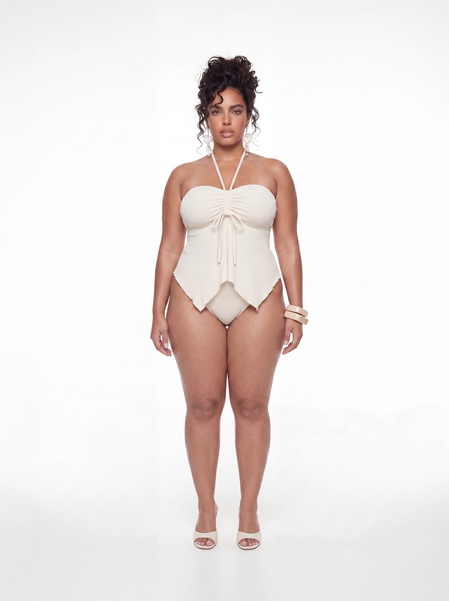 Curve model wearing a cream ribbed one-piece swimsuit featuring a sweetheart neckline, adjustable halter strap, ruched bust with drawstring detail, and pointed handkerchief hem. Styled with statement floral earrings and neutral heels against a white studio background.