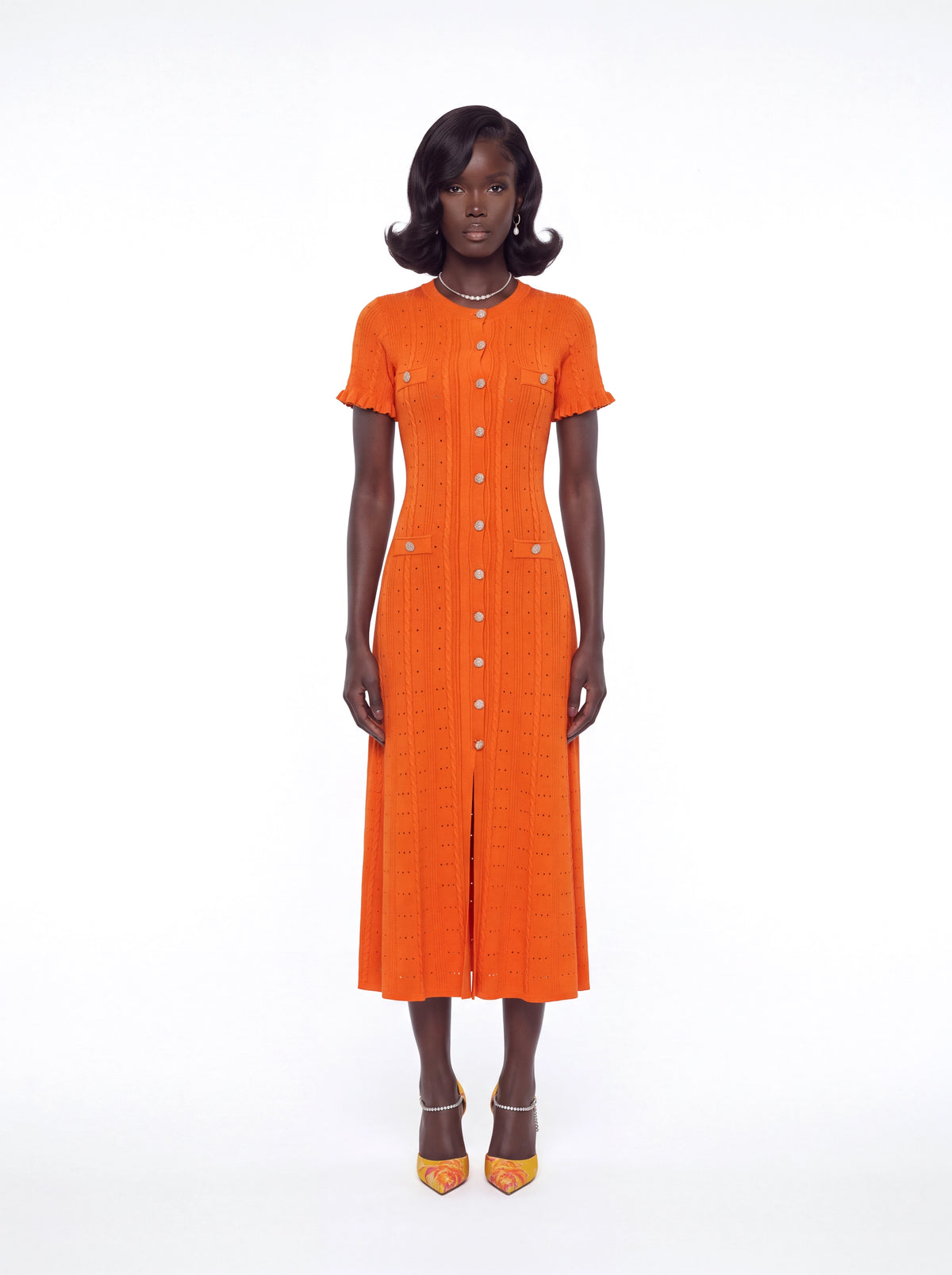 A model stands against a white studio background wearing a bright orange short-sleeve midi dress with a front button placket and front slit, paired with orange and gold pointed-toe heels.