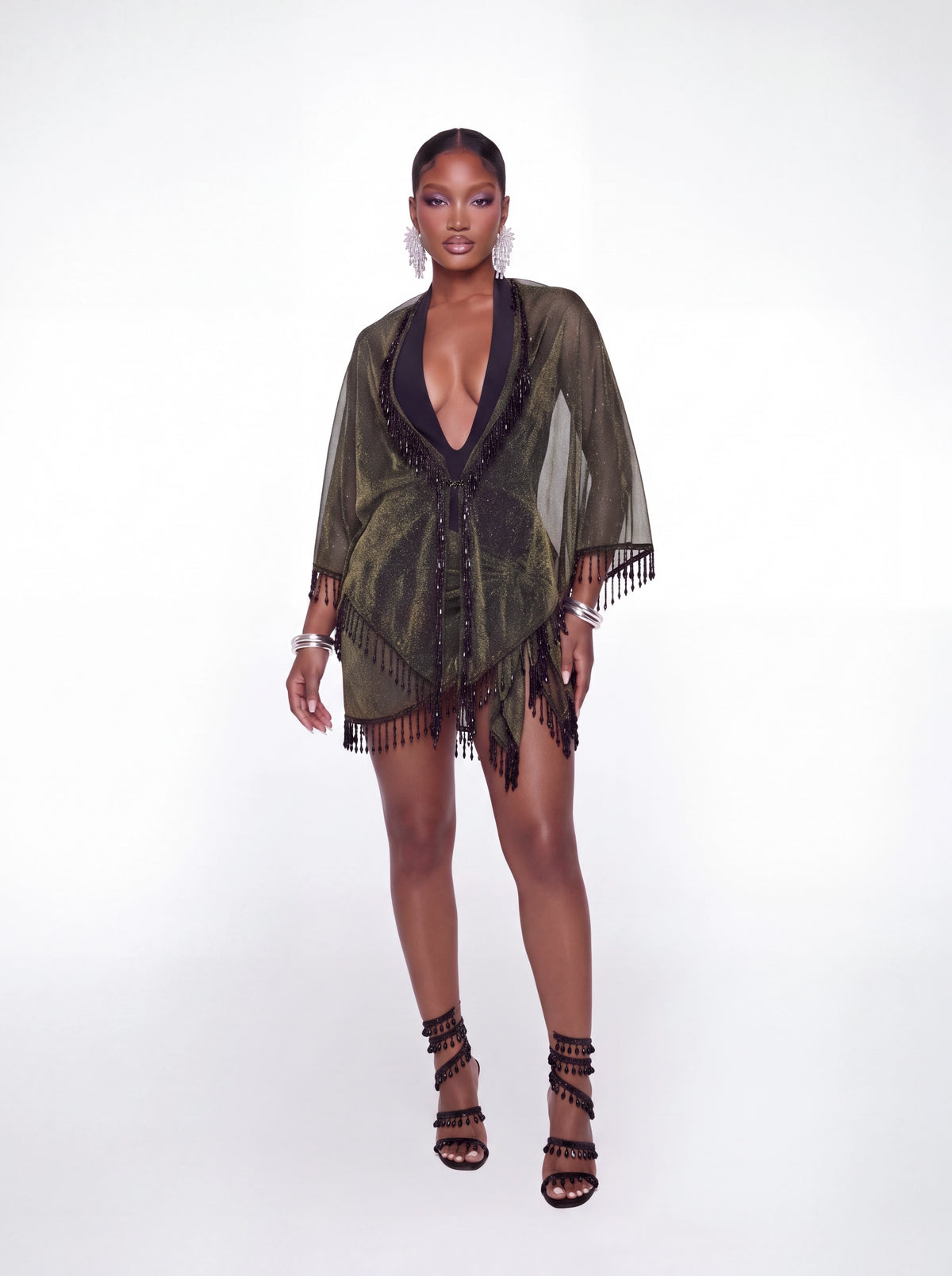 A model stands against a white studio background wearing a sheer olive green fringed cover-up over a black deep V-neck dress, styled with black strappy high-heeled sandals and silver jewelry.