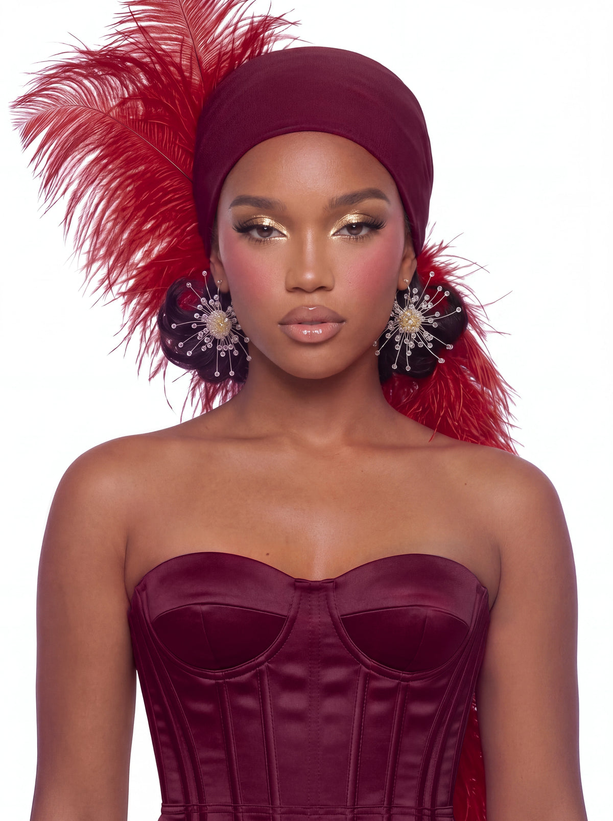 A model wearing a strapless deep burgundy satin corset top, a matching burgundy headband with a large red feather accent, and dramatic statement earrings, set against a solid white background.