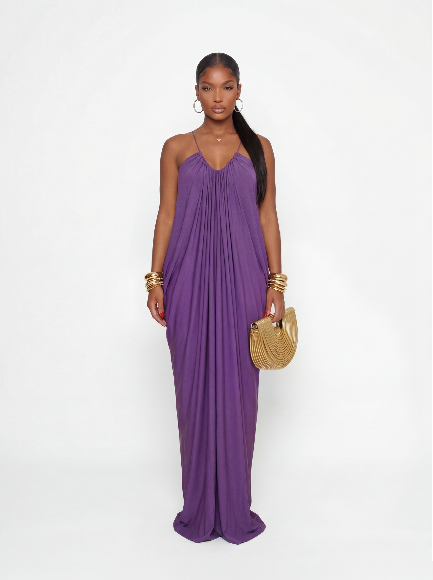 Model wearing a flowy purple maxi dress with spaghetti straps, featuring a pleated draped silhouette, styled with gold bangles and a gold clutch.