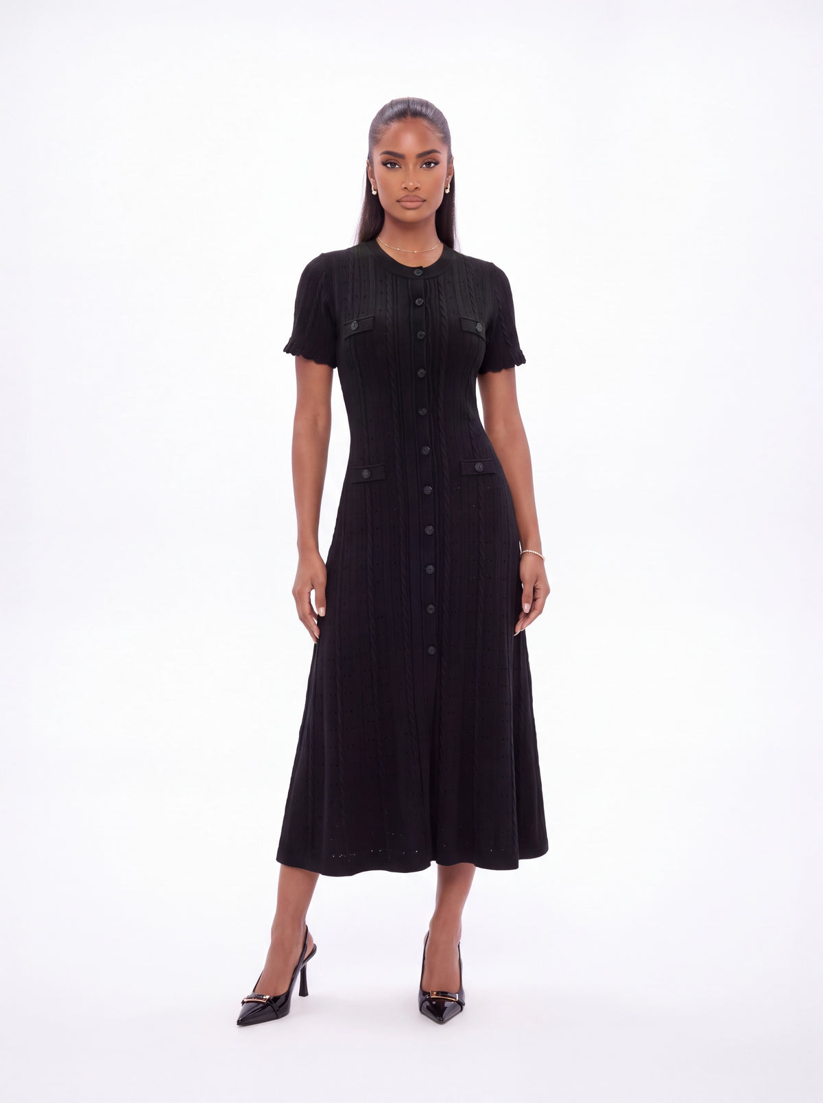 A woman modeling a black short-sleeve midi dress with vertical pleats and a button-front detail, paired with black pointed-toe heels, against a plain white studio background.