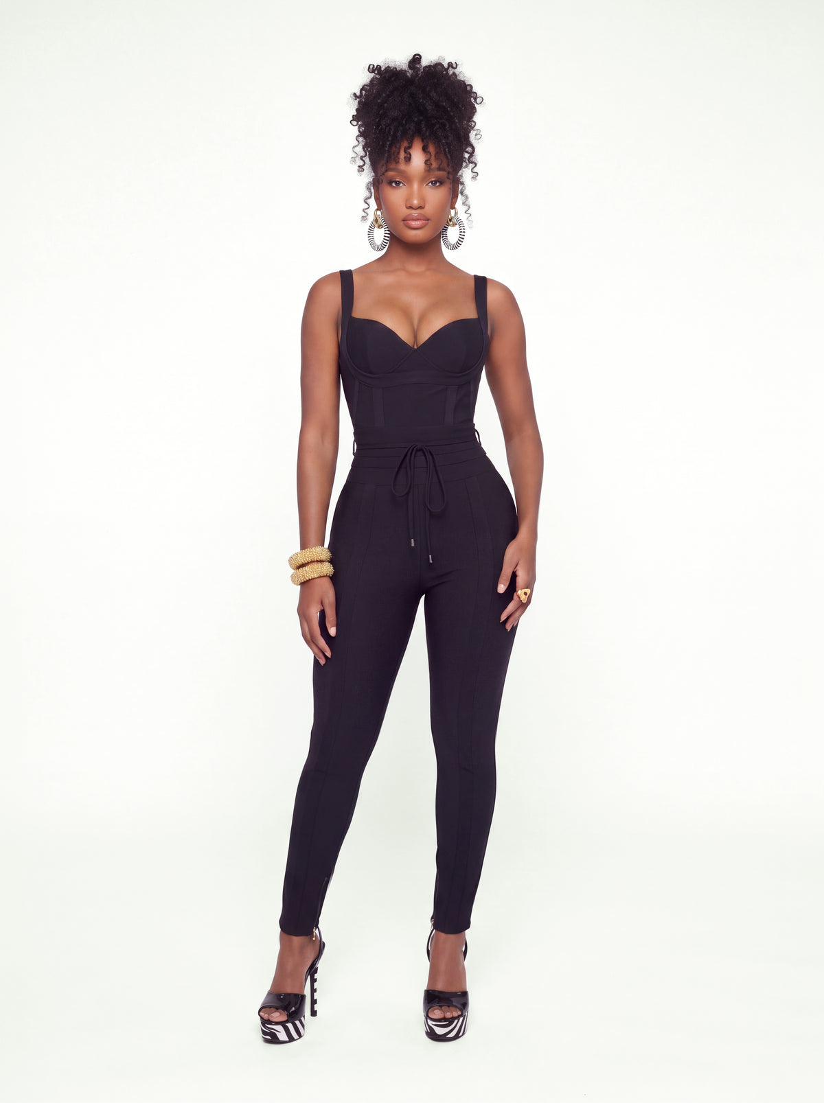Model wearing a black, form-fitting ribbed jumpsuit featuring a structured corset-style bodice, sweetheart neckline, wide shoulder straps, and a waist-defining drawstring. The jumpsuit has slim, tapered legs and is styled with gold accessories and heels.