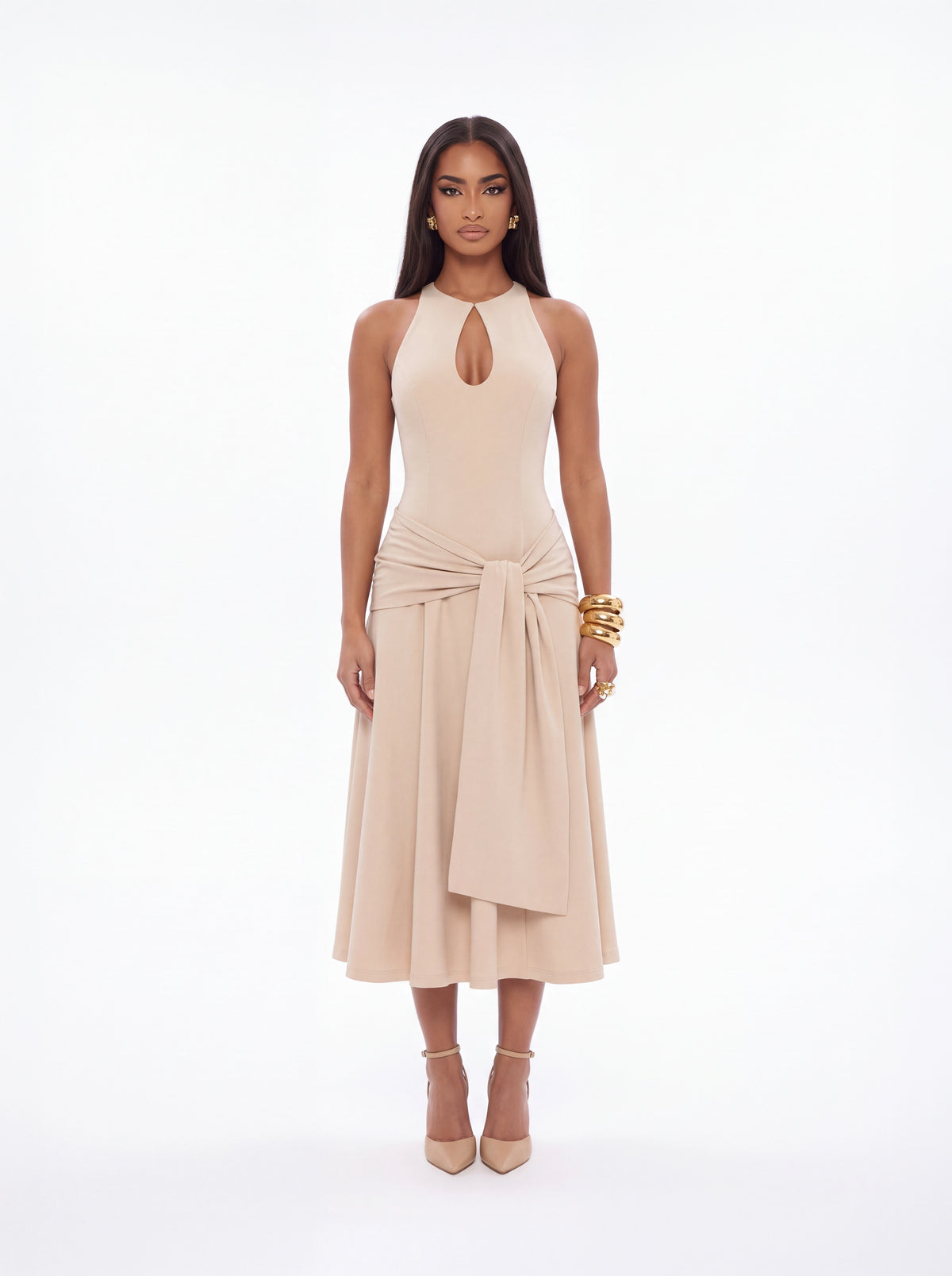 A model stands against a seamless white studio background wearing a sleeveless beige midi dress with a keyhole neckline, a knotted waist, and a draped skirt. She is wearing nude high heels and gold bracelets.
