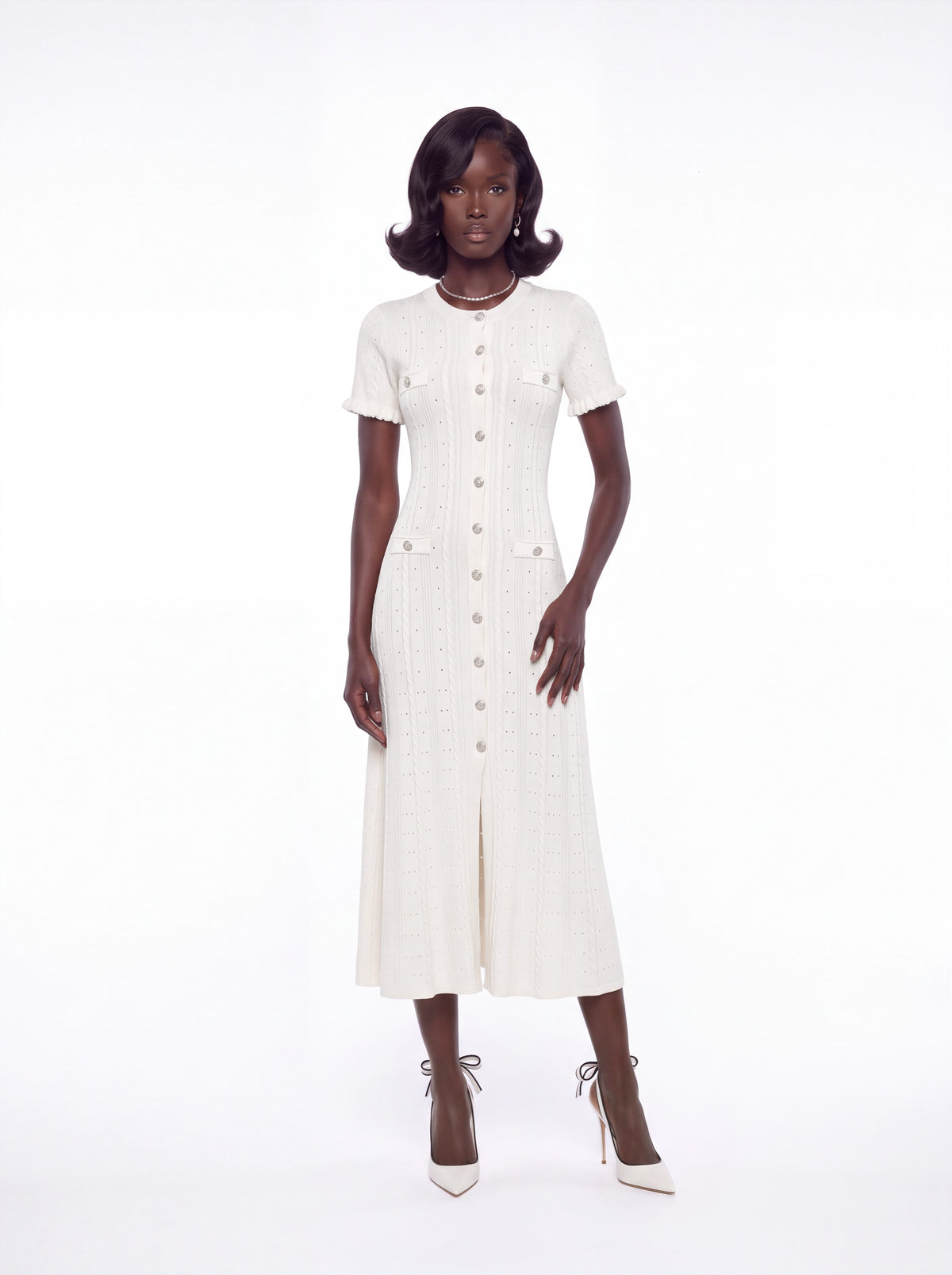 A model stands against a seamless white studio background wearing a short-sleeved cream-colored textured midi dress with a button-down front and matching cream pointed-toe heels.