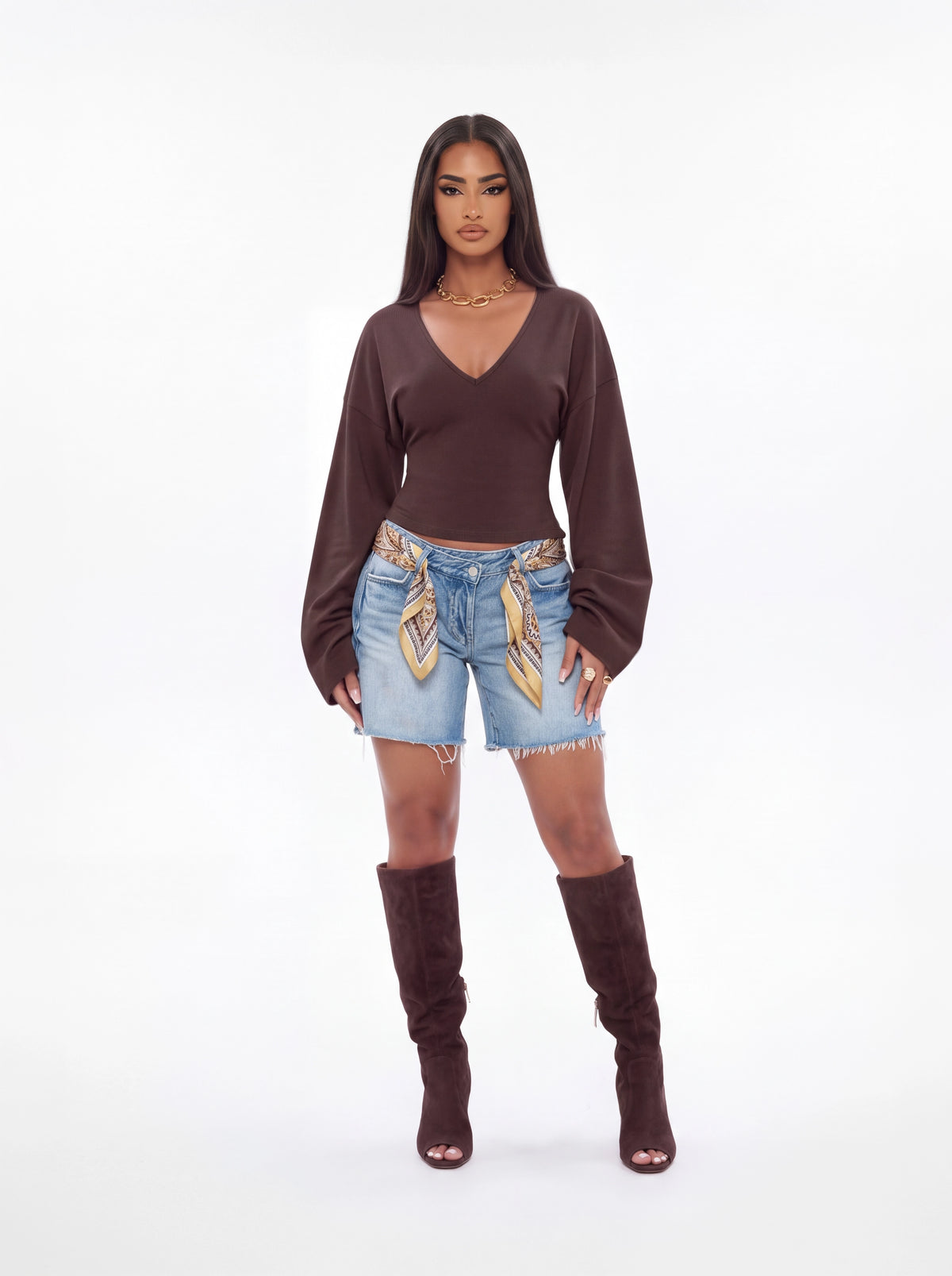 A model stands against a white studio background wearing a dark brown long-sleeve cropped top, light blue denim shorts with gold patterned embroidery, and matching dark brown heeled boots.